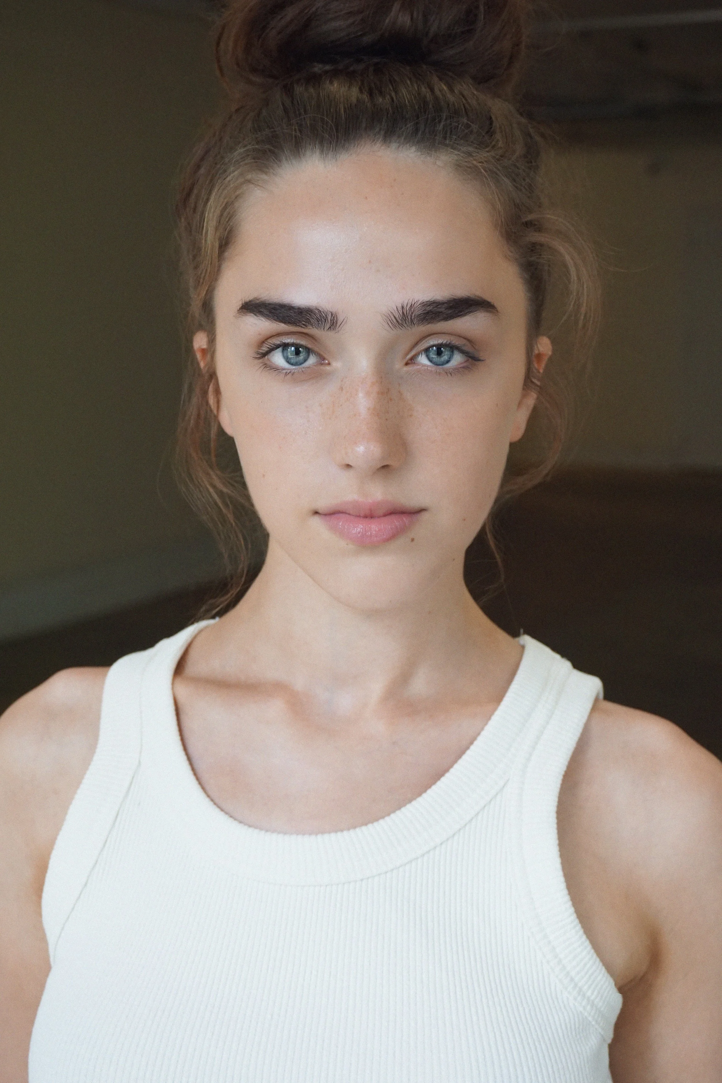 Close-up portrait of a young woman with blue eyes, brown hair styled in a high bun, wearing a white sleeveless top, looking directly at the camera.