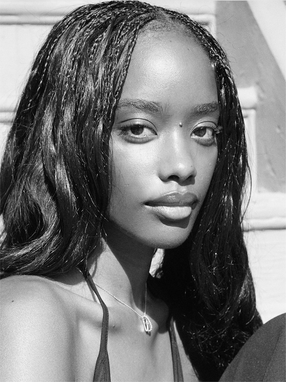Close-up black and white portrait of a young woman with long wavy hair, wearing a necklace with a 'D' pendant, looking directly at the camera.