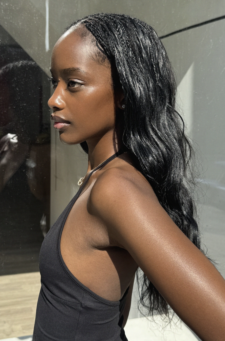A young Black woman with long, wavy black hair, wearing a black sleeveless top, looking to her left, with a reflective surface and indoor setting in the background.