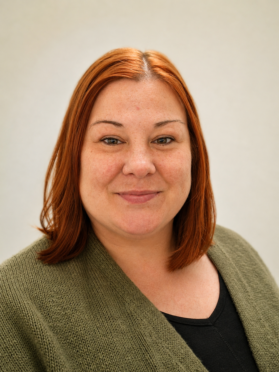A headshot of a woman with shoulder-length red hair, fair skin, and freckles, wearing a green sweater over a black top, smiling slightly with a plain light background.