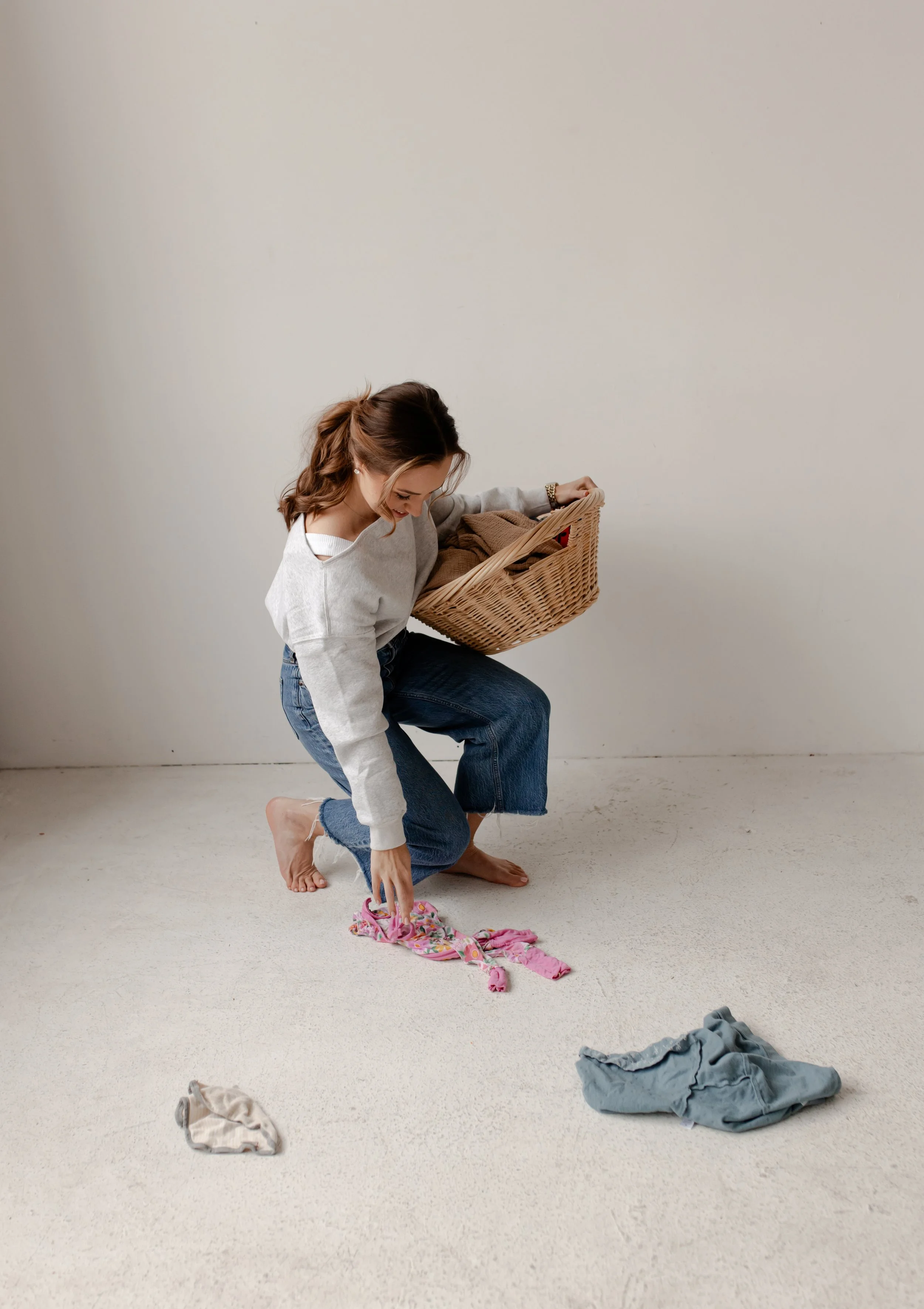 A woman kneeling on a beige carpeted floor, wearing a gray sweater and jeans, placing a pink piece of clothing on the ground. She holds a laundry basket filled with folded clothes. Several unfolded clothes are scattered on the floor around her, and the background features a plain, cream-colored wall.