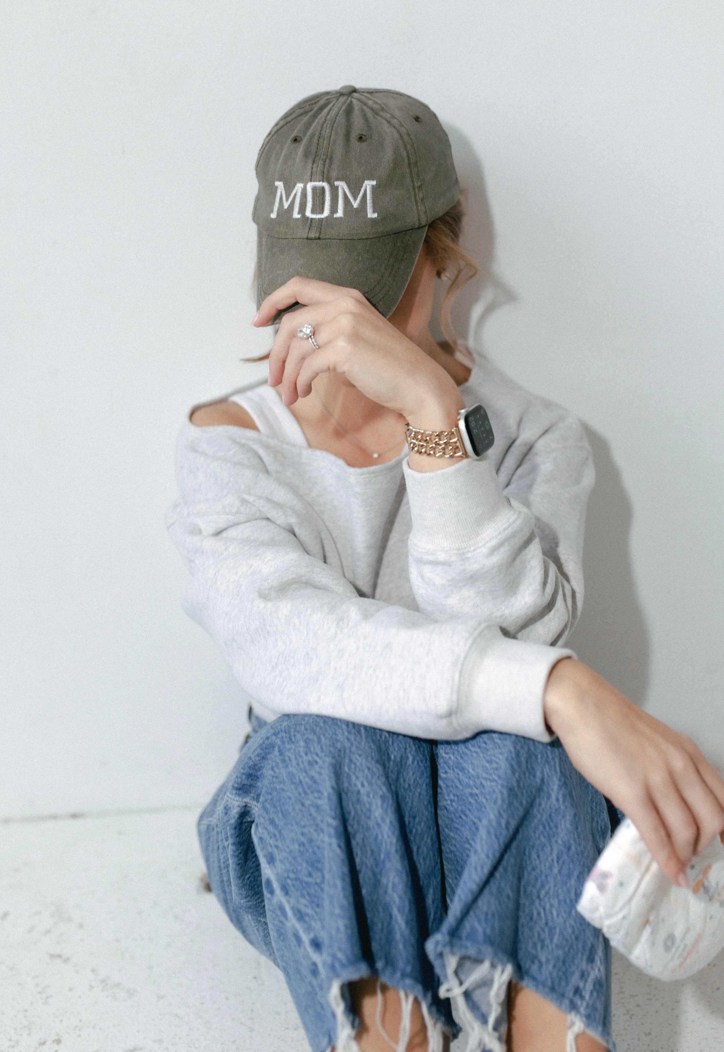 Person sitting on the floor wearing a baseball cap that says 'MOM', casual gray sweatshirt with a shoulder cutout, ripped jeans, a smartwatch, and a ring, holding a snack bag.