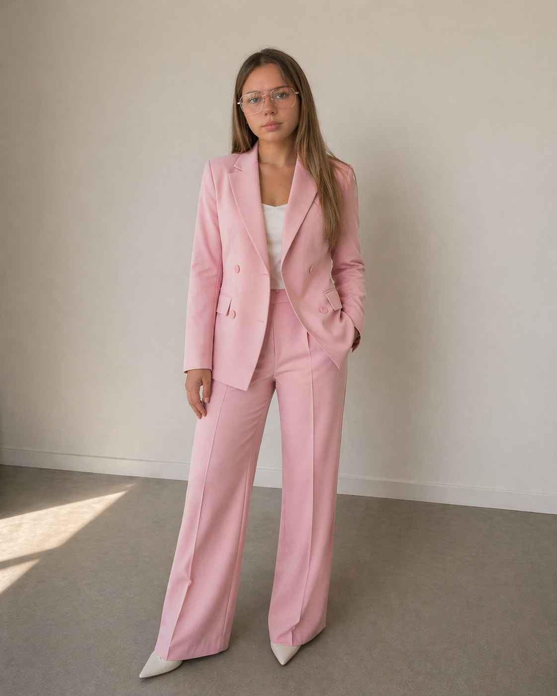 Woman in a pink pantsuit, white blouse, and white heels standing indoors against a plain wall.