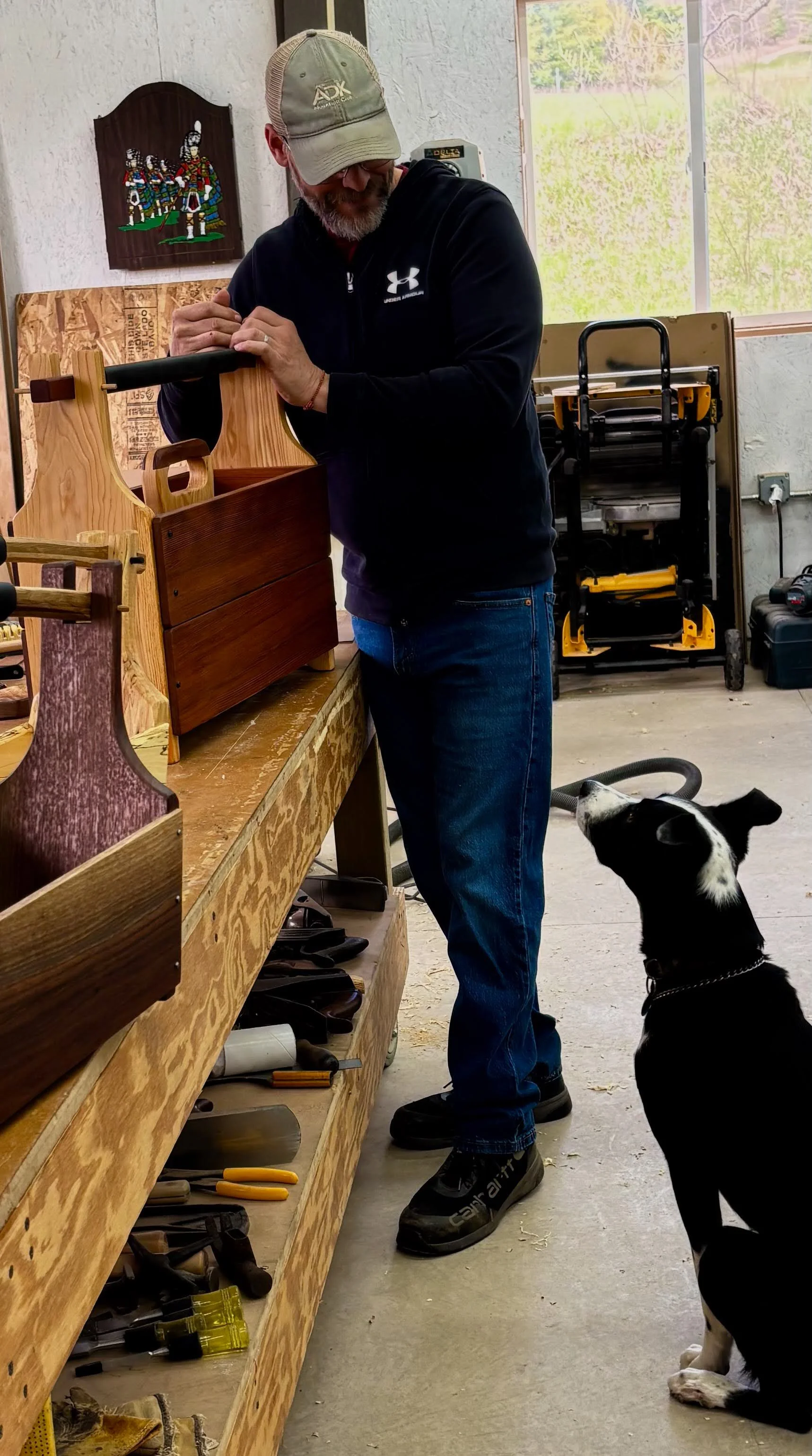 Craftsman Tim Pierce working on woodworking tools on a workbench, with a black and white dog sitting beside him in a workshop.