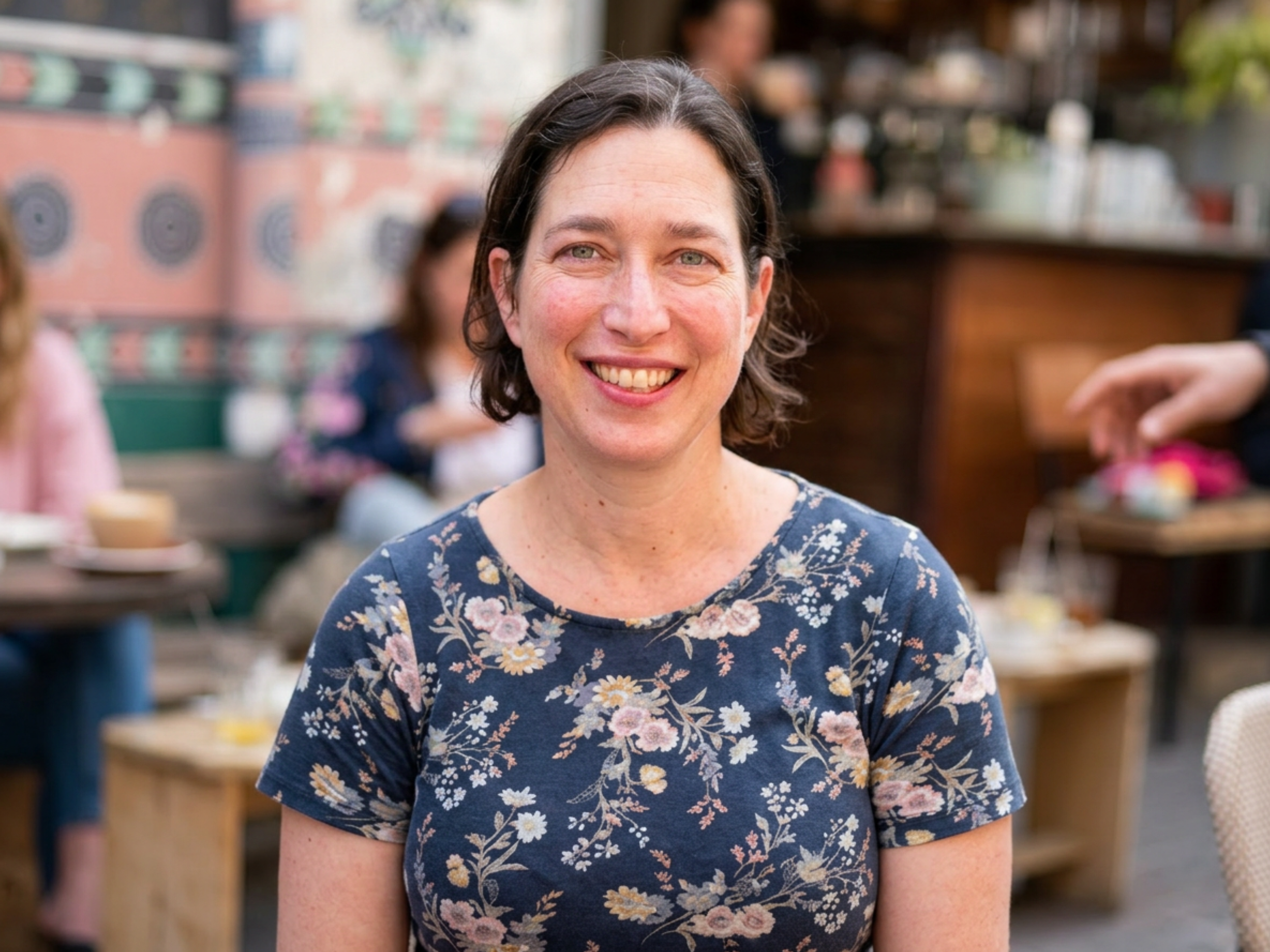 Irit's headshot: a woman with short brown hair, wearing a blue floral shirt and smiling, sitting in an outdoor setting that appears to be a cafe with blurred background of tables.