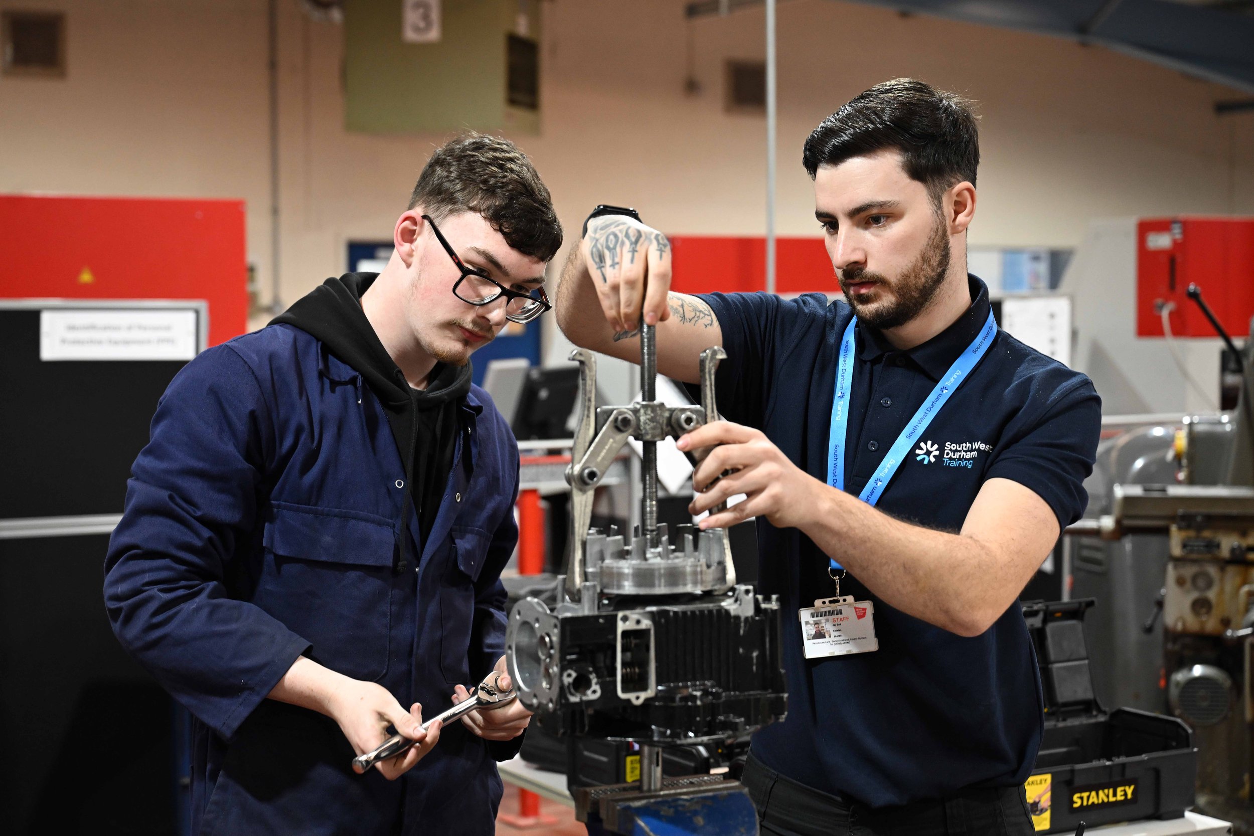 Two men working on machinery in an industrial setting, one holding a wrench and the other adjusting a part with a tool, both wearing navy uniforms.