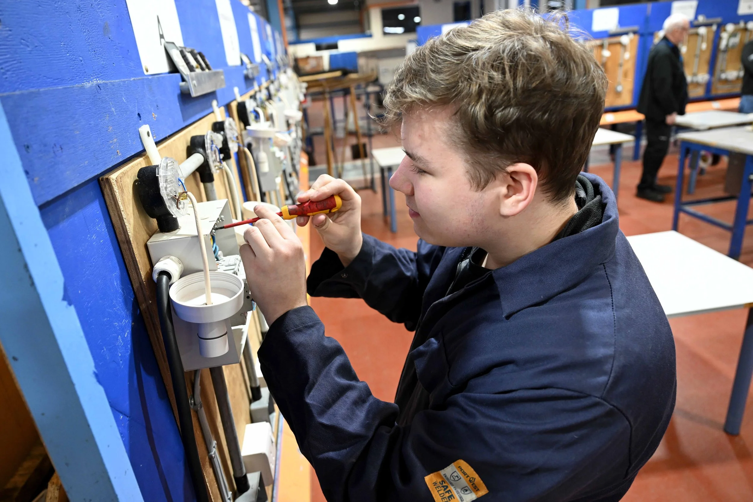 A young man working on an electrical project at a blue workbench, using a screwdriver and surrounded by electrical components and sockets in a workshop or classroom setting.