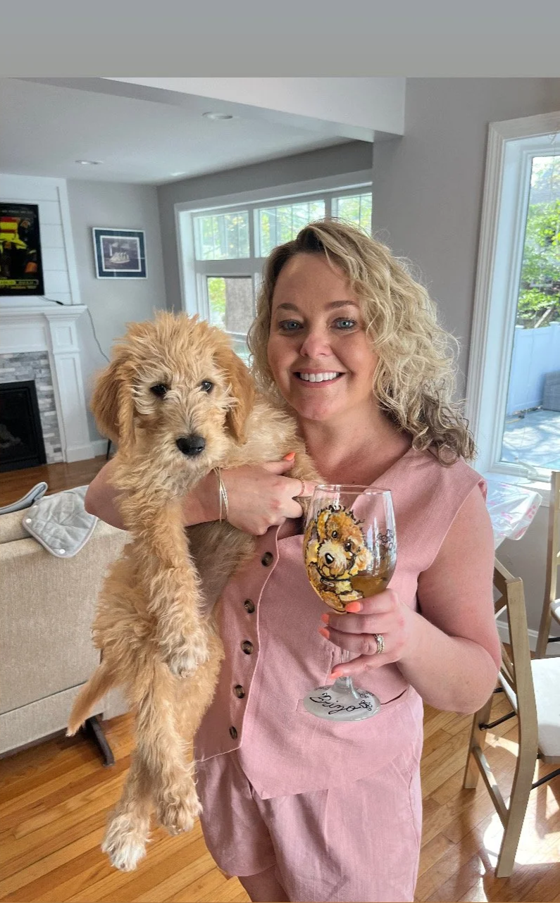A woman holding a puppy in a living room, smiling and holding a glass of wine with a lion cartoon on it, with large windows and a fireplace in the background.