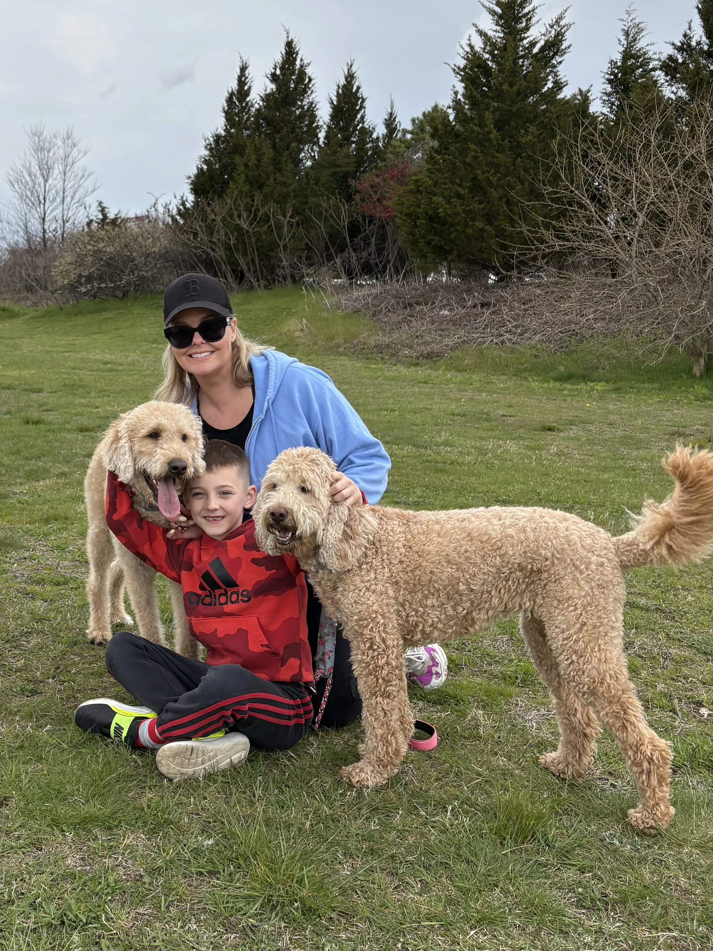 Woman wearing sunglasses, a black cap, and a blue hoodie, kneeling on grass, smiling with a boy in a red camouflage hoodie, surrounded by several large, fluffy, tan-colored dogs outdoors near trees.