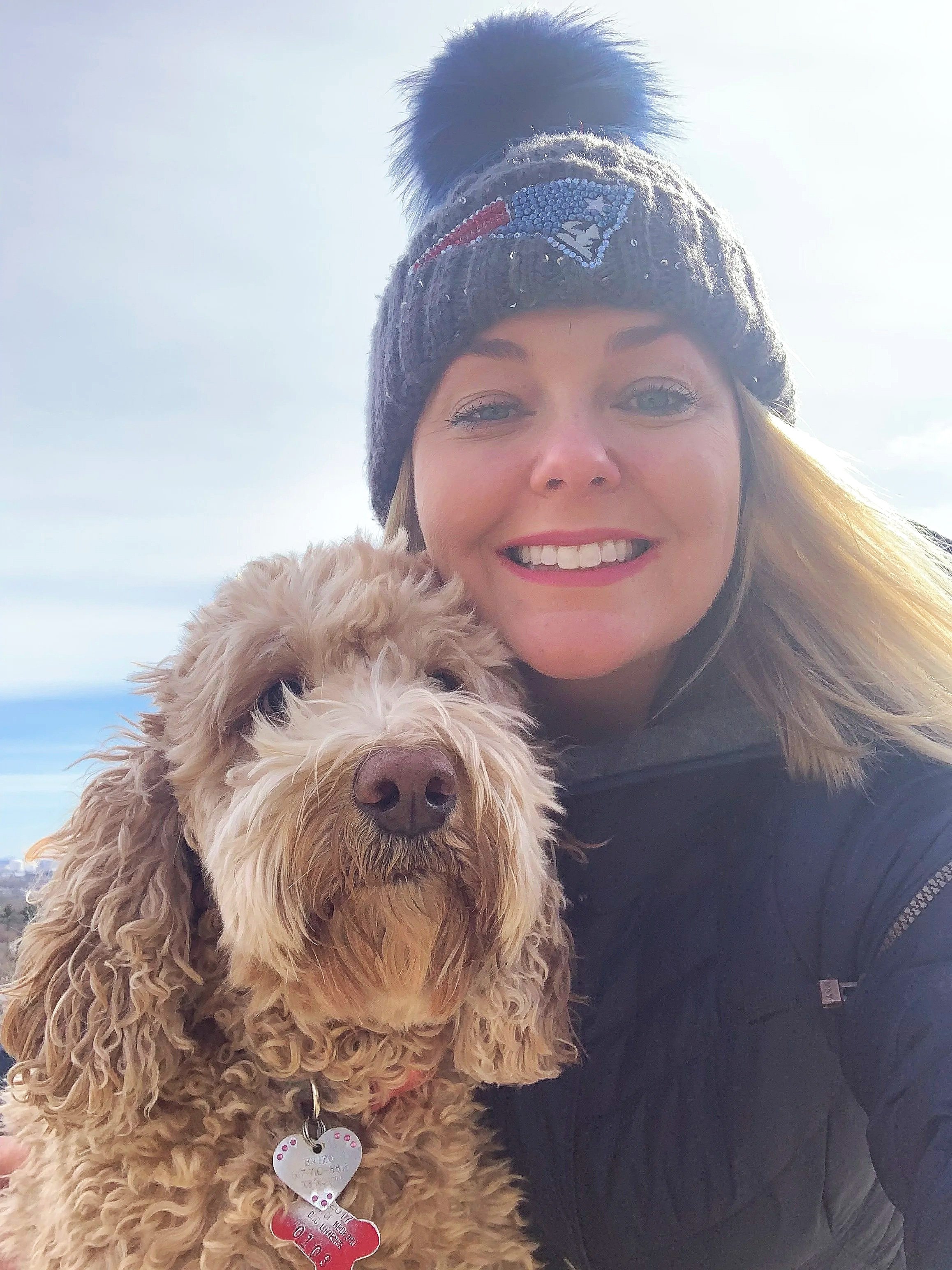 A woman is smiling for a close-up selfie with her dog during daytime. She is wearing a knit hat with a pom-pom on top, and the dog has a fur coat with a tag. The background shows an outdoor setting with a cloudy sky.