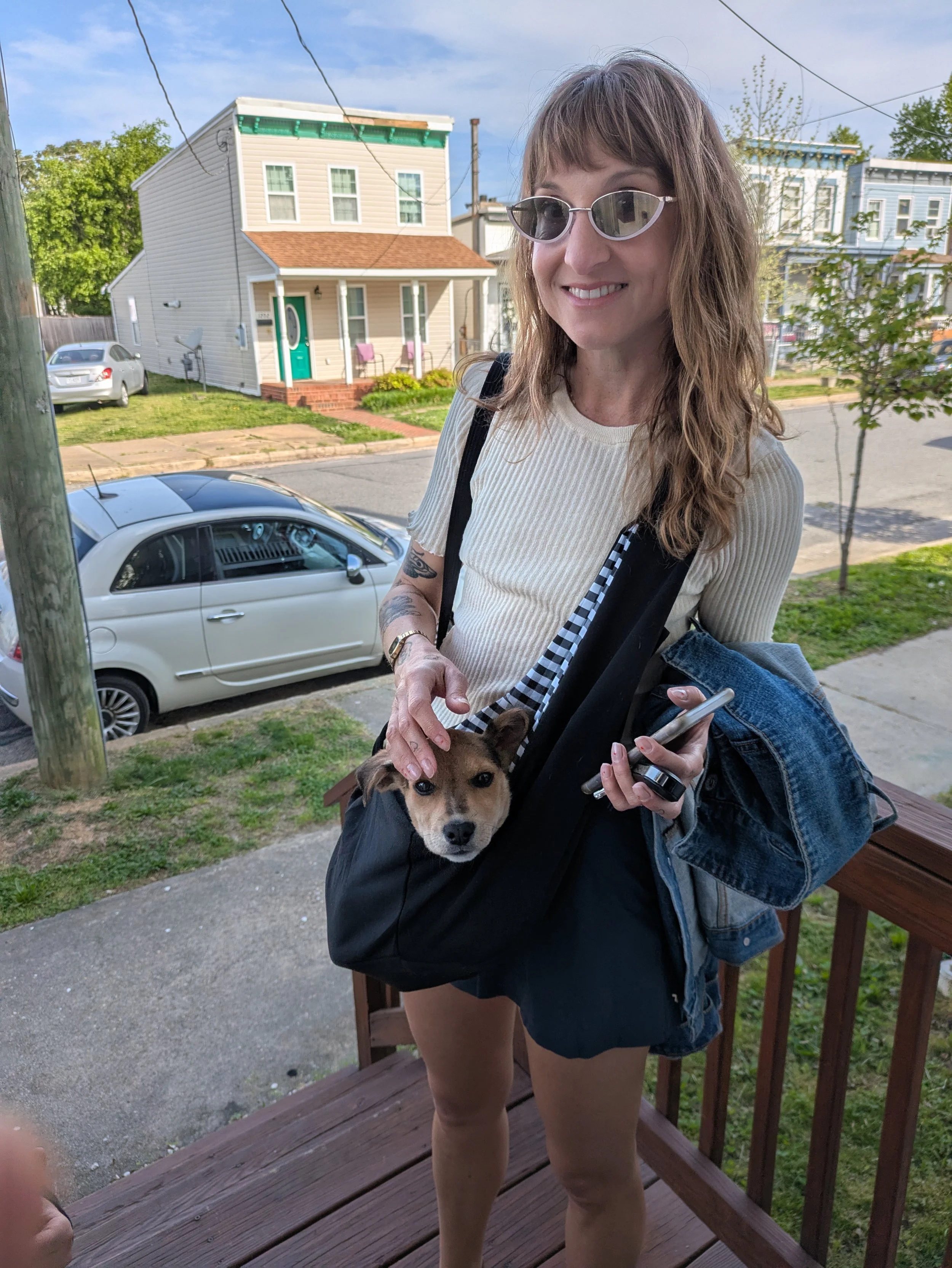 A woman with wavy brown hair wearing sunglasses, a cream-colored ribbed shirt, and denim shorts, holding a small brown and white puppy, standing on a wooden deck in a residential neighborhood with cars parked on the street and houses in the background.