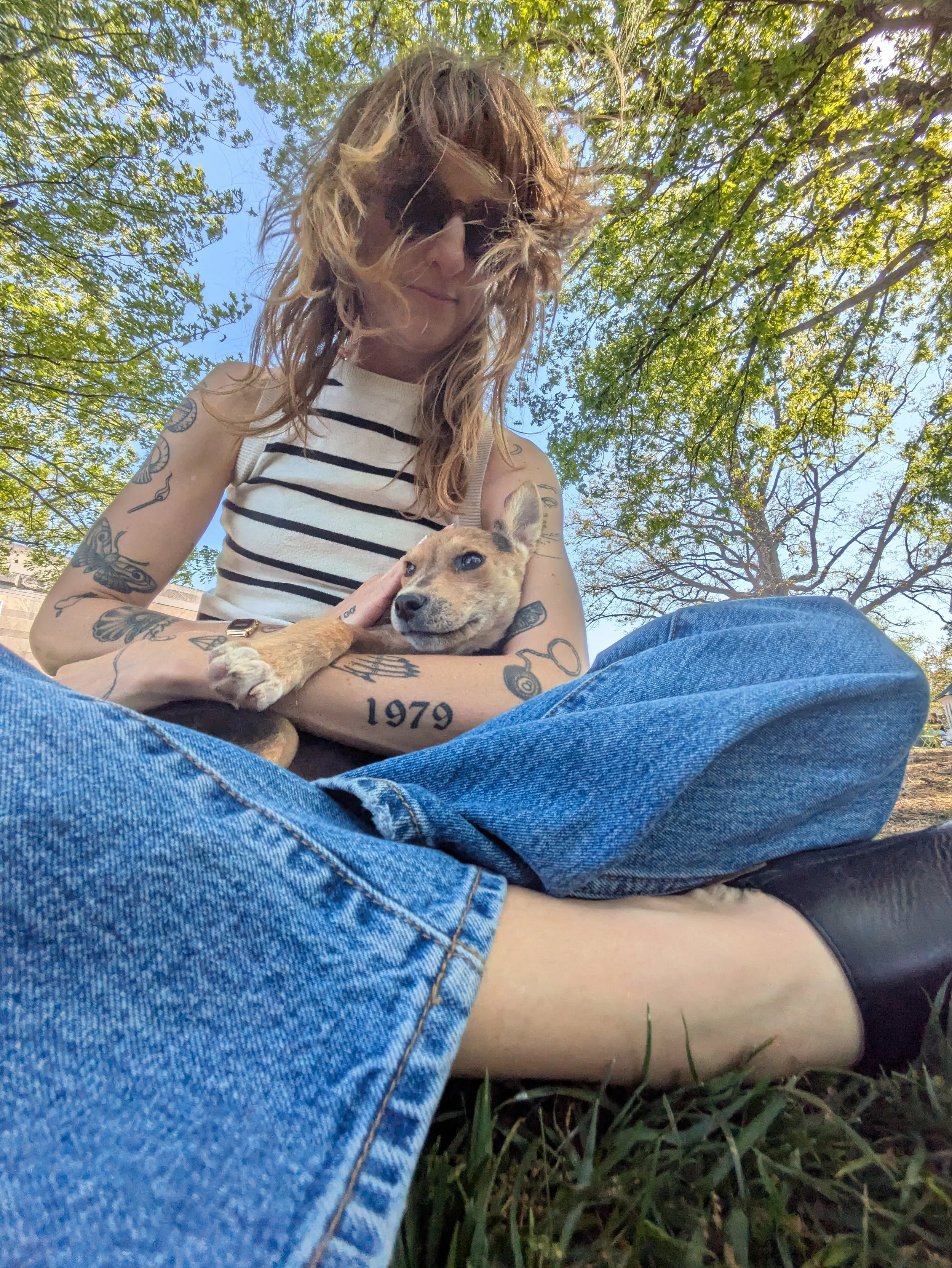 A woman with tattoos on her arms, wearing sunglasses and a striped sleeveless top, is sitting outdoors on grass, holding a small puppy. The background shows trees with green leaves and a blue sky.