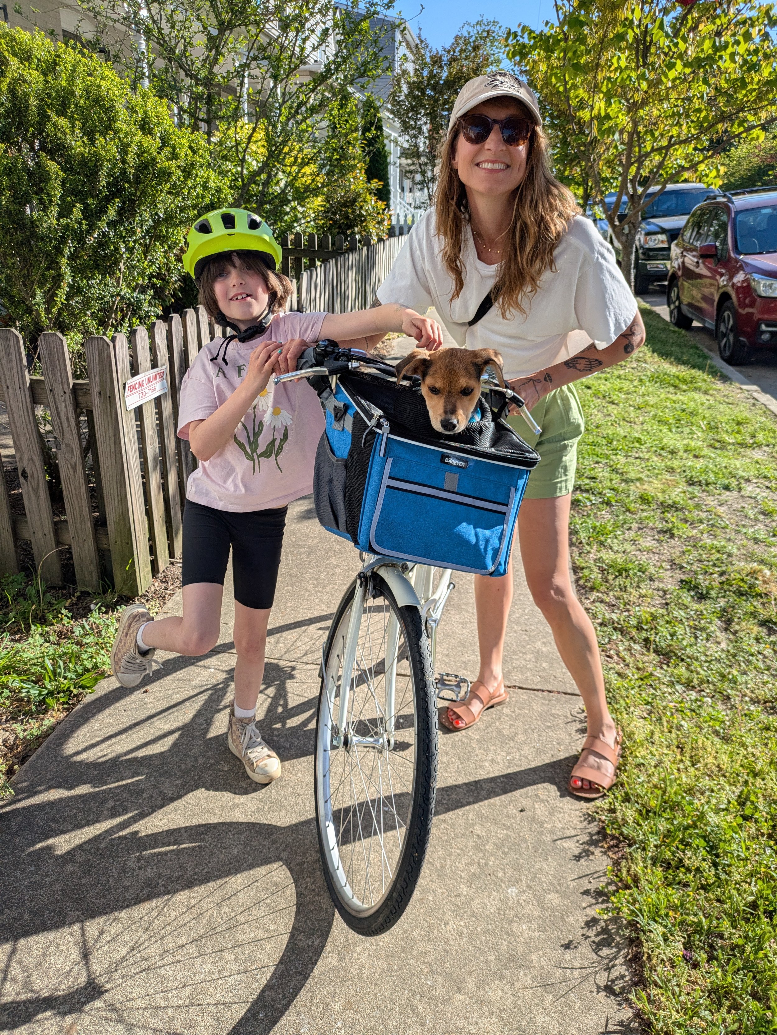 A woman with long hair, sunglasses, and a white hat smiling while standing next to a young girl with short hair, a green bike helmet, and casual clothing, holding a yellow and brown puppy in a front bike basket, outdoors on a sidewalk with green bushes, trees, and parked cars in the background.