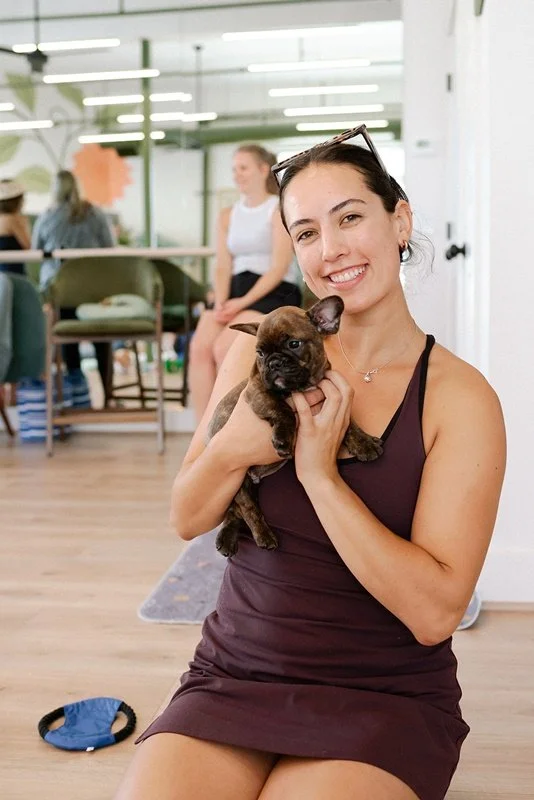 woman in a sleeveless purple dress sitting on the floor, smiling, holding a small brindle French Bulldog puppy indoors, with people and furniture in the background.