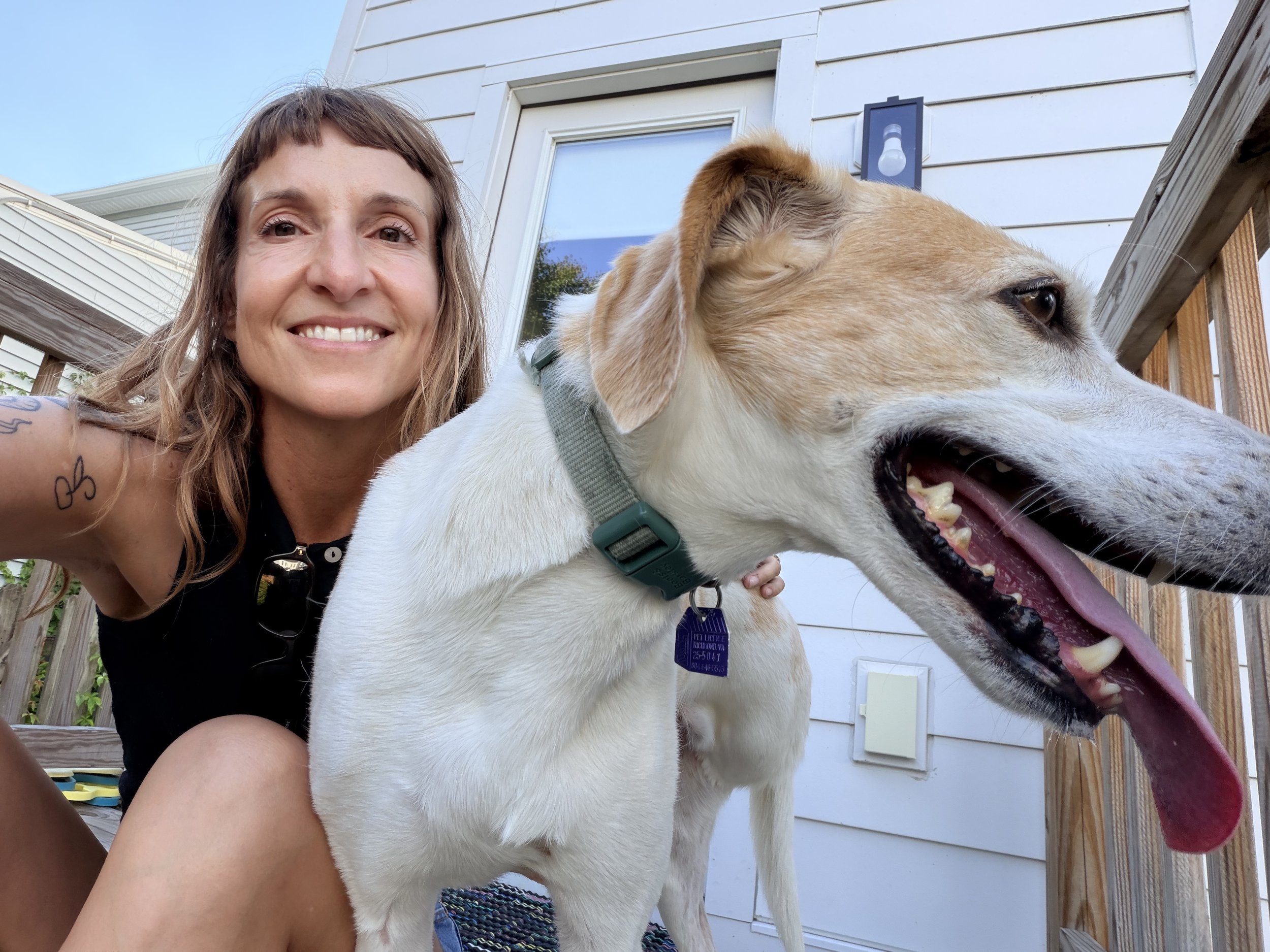 A woman with wavy brown hair smiling at the camera, standing next to a large light-colored dog with a green collar, on a wooden deck outside a house with white siding.