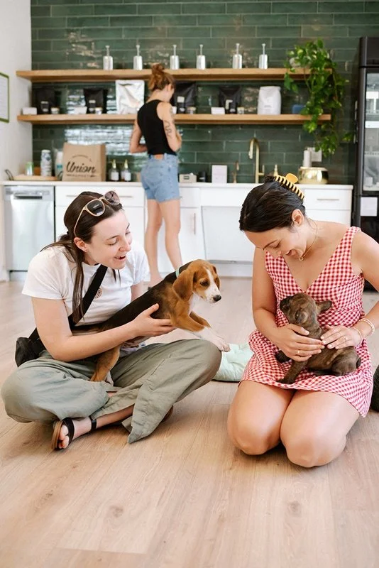 Two women sitting on the floor in a studio, each holding a dog, with a woman in the background browsing shelves.