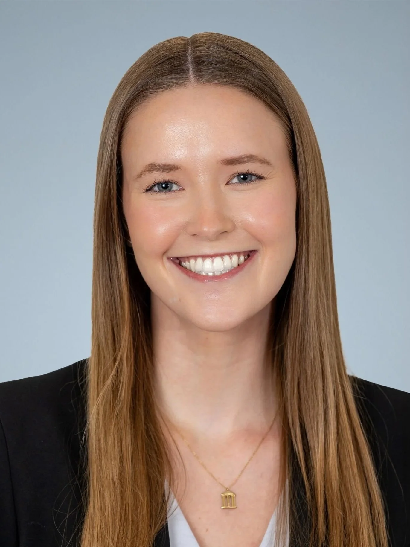 Portrait of a woman with long, straight, light brown hair, smiling, wearing a black blazer, white top, and a gold necklace with a small pendant.