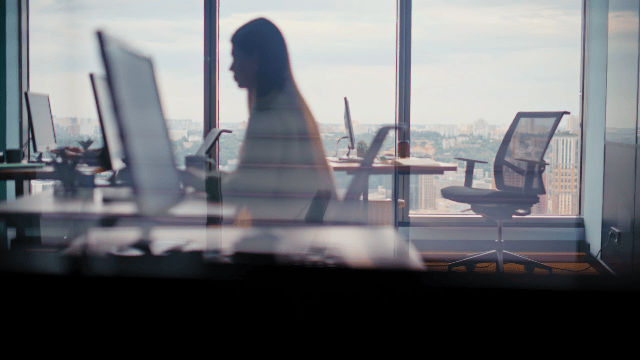 Silhouette of a woman working at a desk in a modern office with large windows overlooking a city skyline.