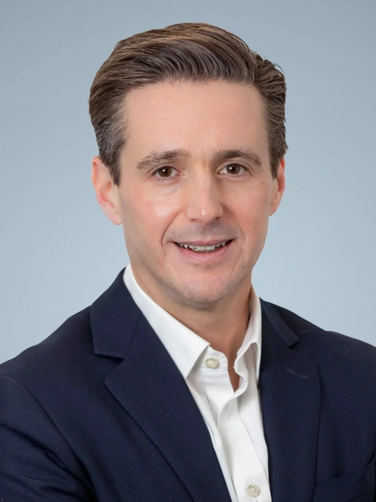 A man with brown hair in a professional suit smiles at the camera against a light blue background.