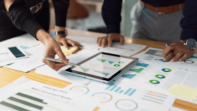 People analyze financial charts and data on a wooden table using a tablet, sheets of paper, and pens in an office setting.