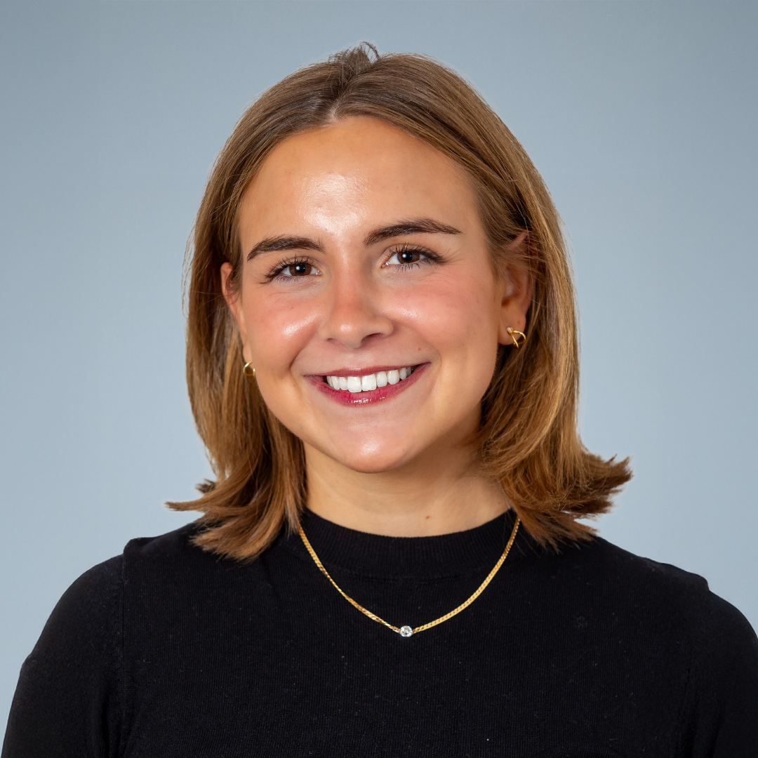 A woman with shoulder-length light brown hair, smiling, wearing a black top, gold jewelry, and a light blue background.