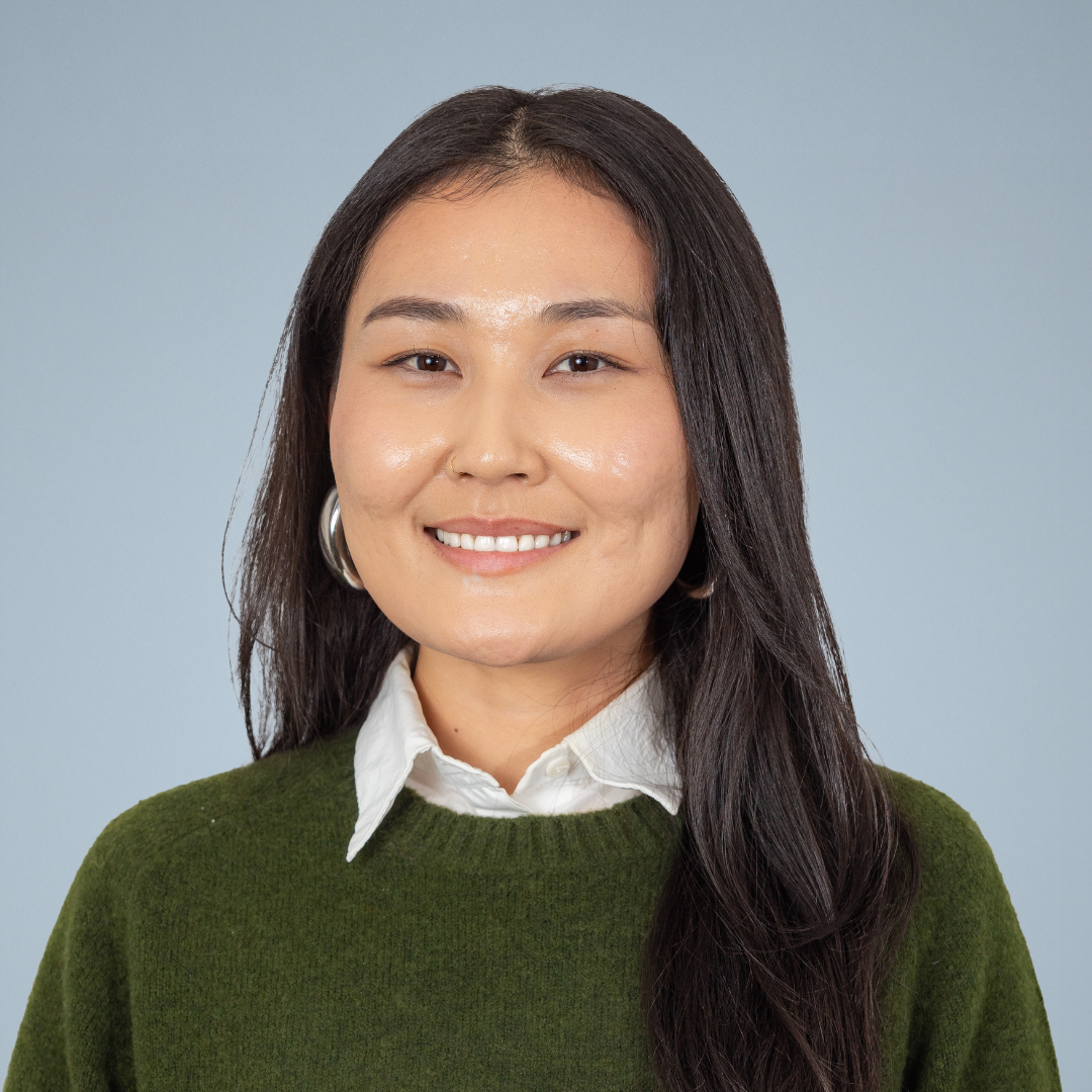 A young woman with long dark hair, smiling, wearing a white collared shirt under a green sweater, against a light blue background.