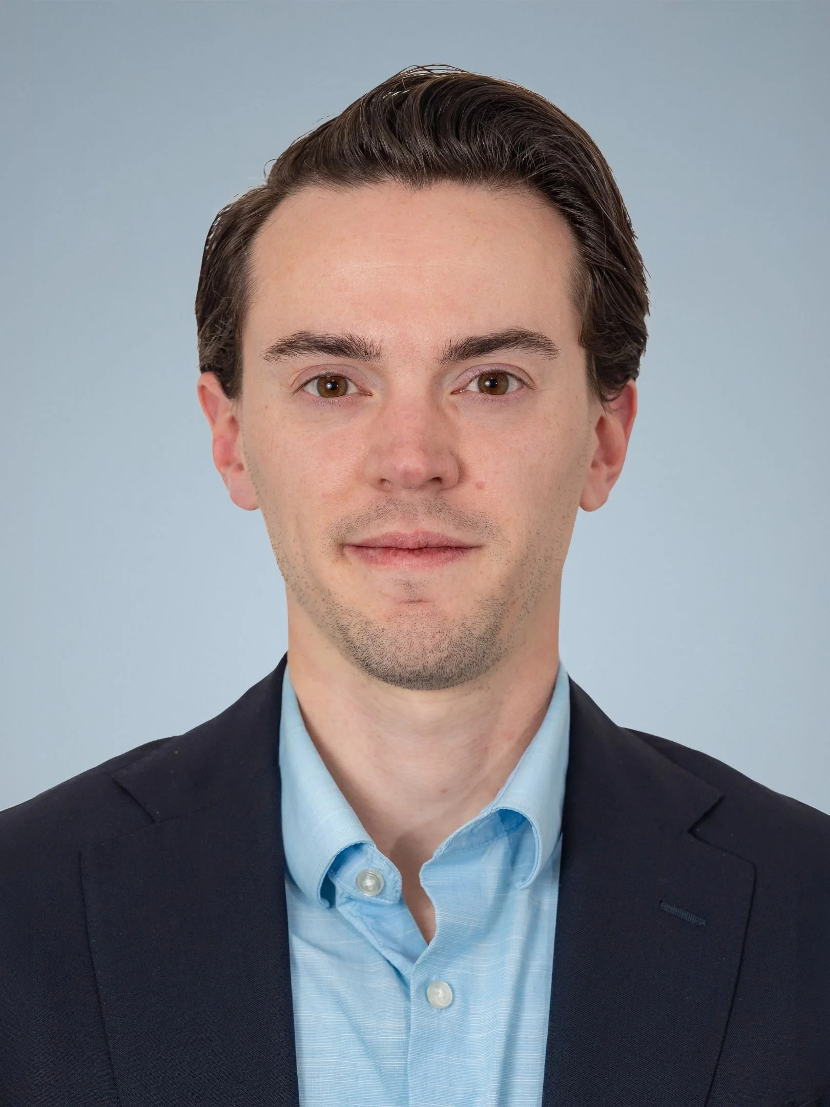 Head and shoulders portrait of a young man with dark brown hair, wearing a navy blazer and a light blue dress shirt, against a plain light blue background.