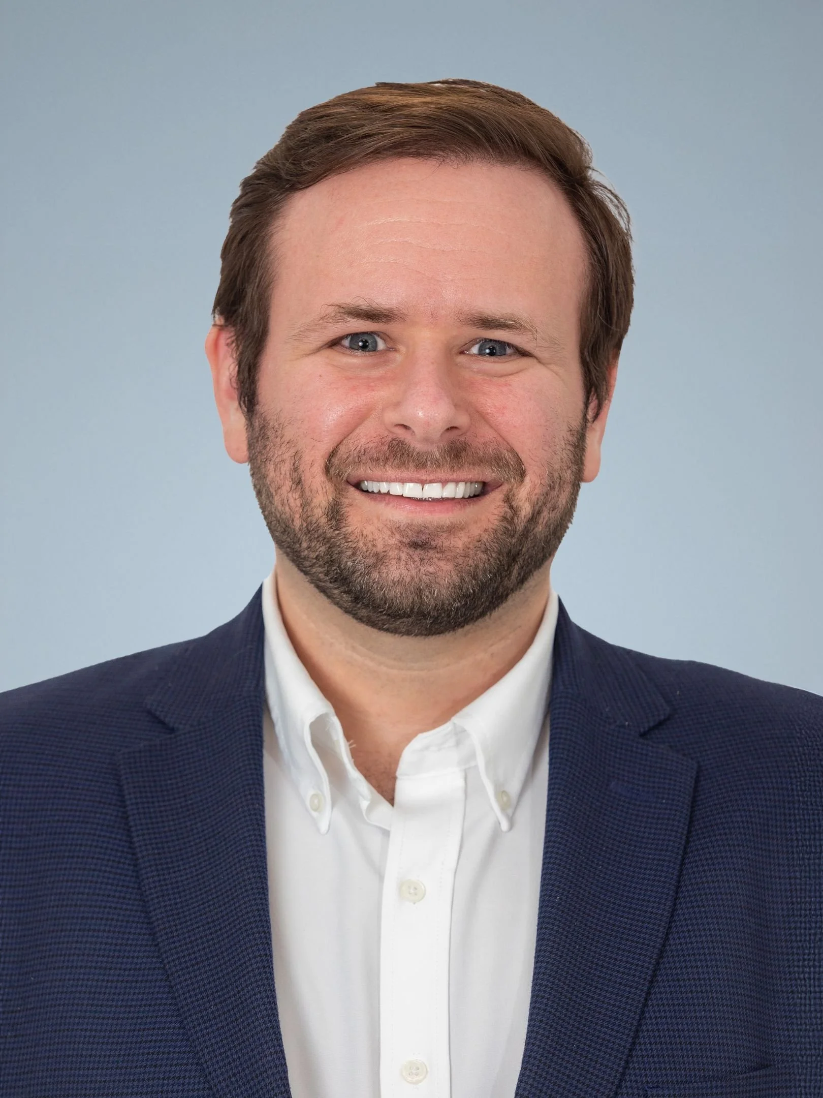 Headshot of a man with light brown hair and beard, wearing a navy blazer and white shirt, smiling against a light blue background.