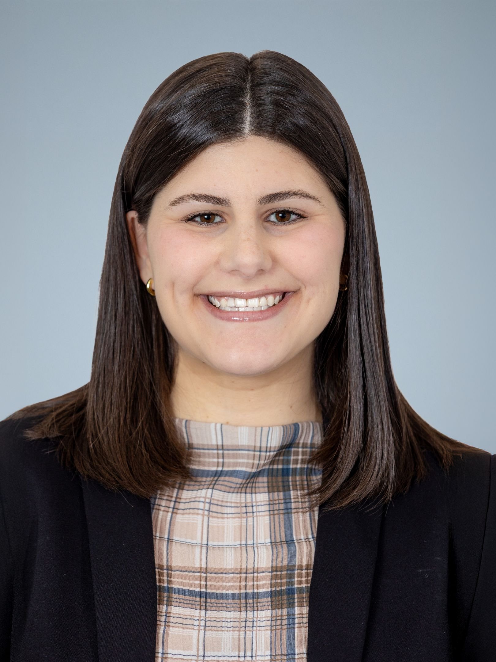 A woman with straight brown hair smiling at the camera, wearing a black blazer over a plaid top, with a light blue background.