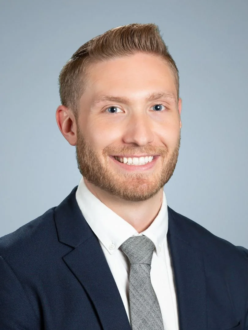 A young man with light brown hair, a beard, and blue eyes wearing a dark suit, white shirt, and gray tie, smiling against a light gray background.