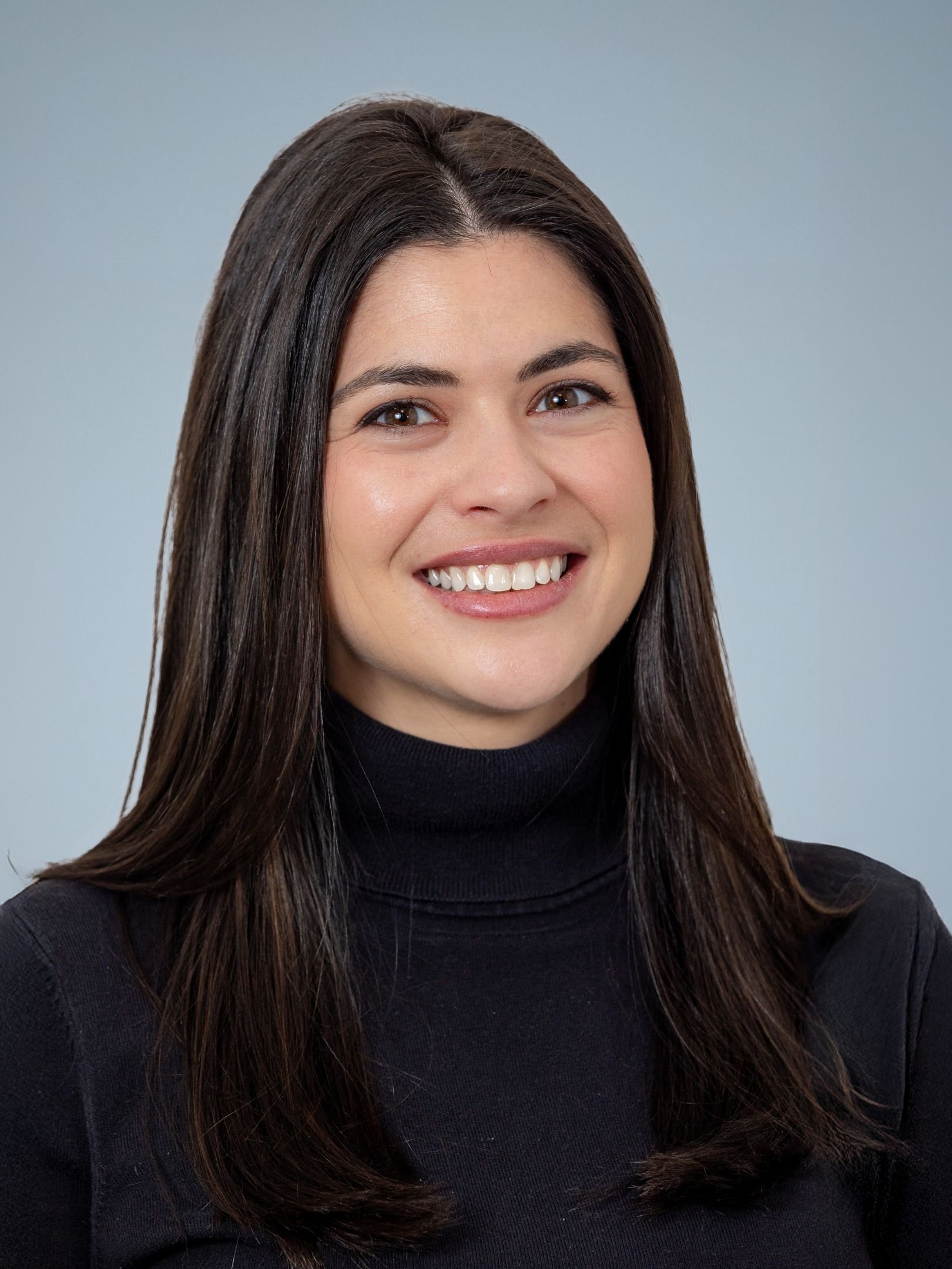 A smiling woman with long dark brown hair, wearing a black turtleneck, against a light grey background.