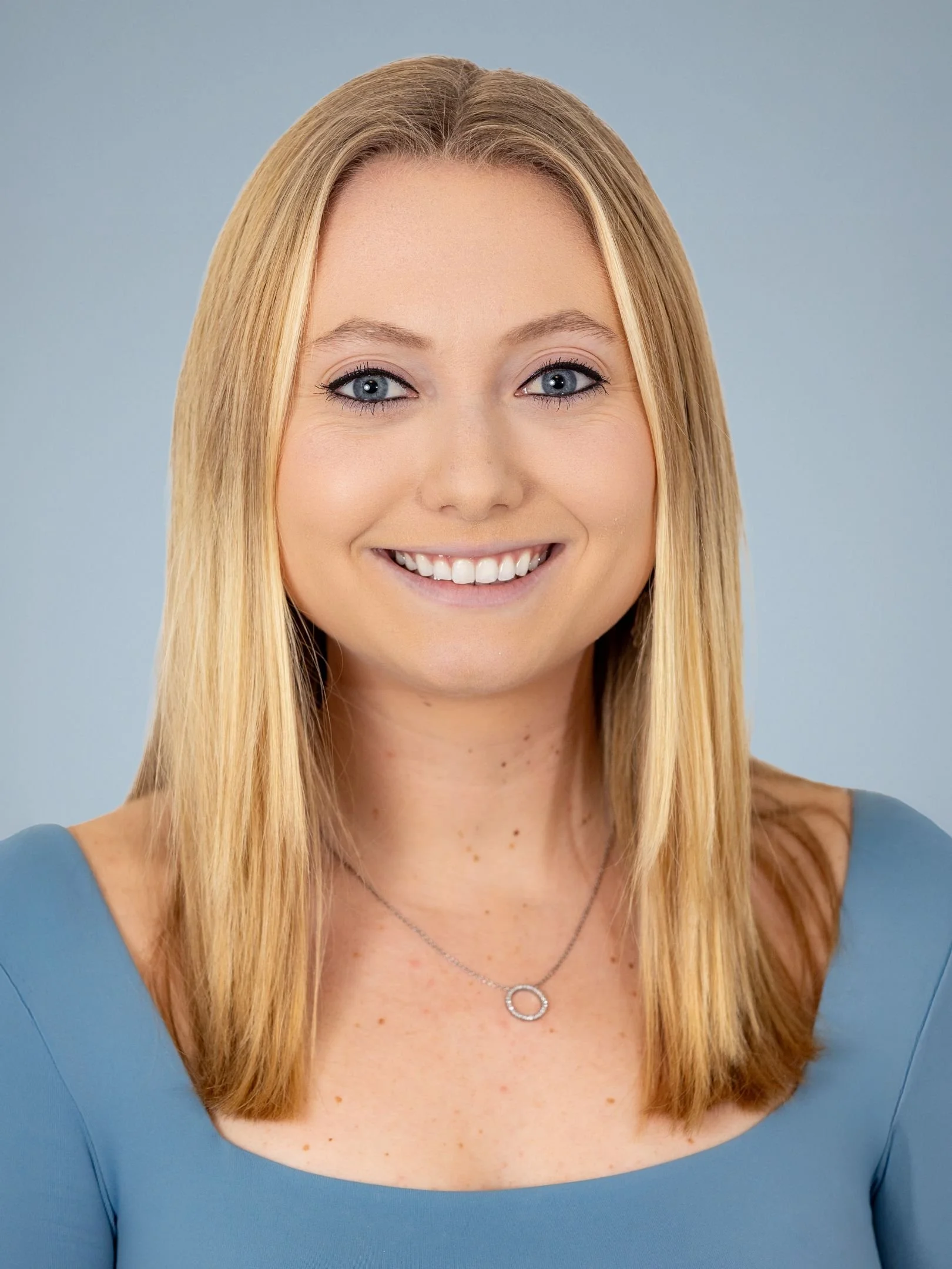 A smiling woman with blue eyes, blonde hair, wearing a blue top and a silver necklace.