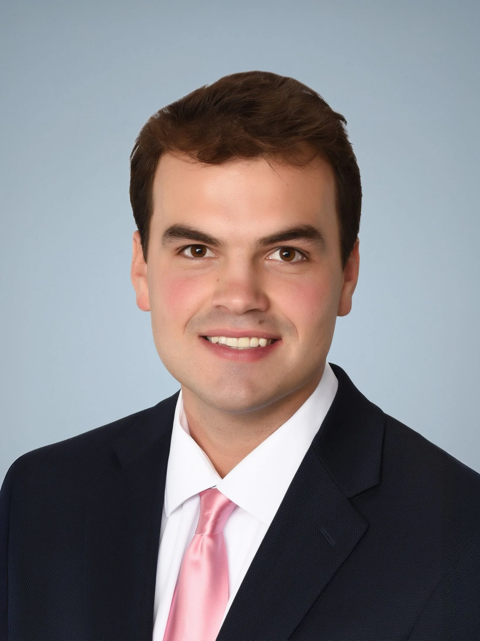 A young man with brown hair smiling in a business suit and pink tie, against a light blue background.