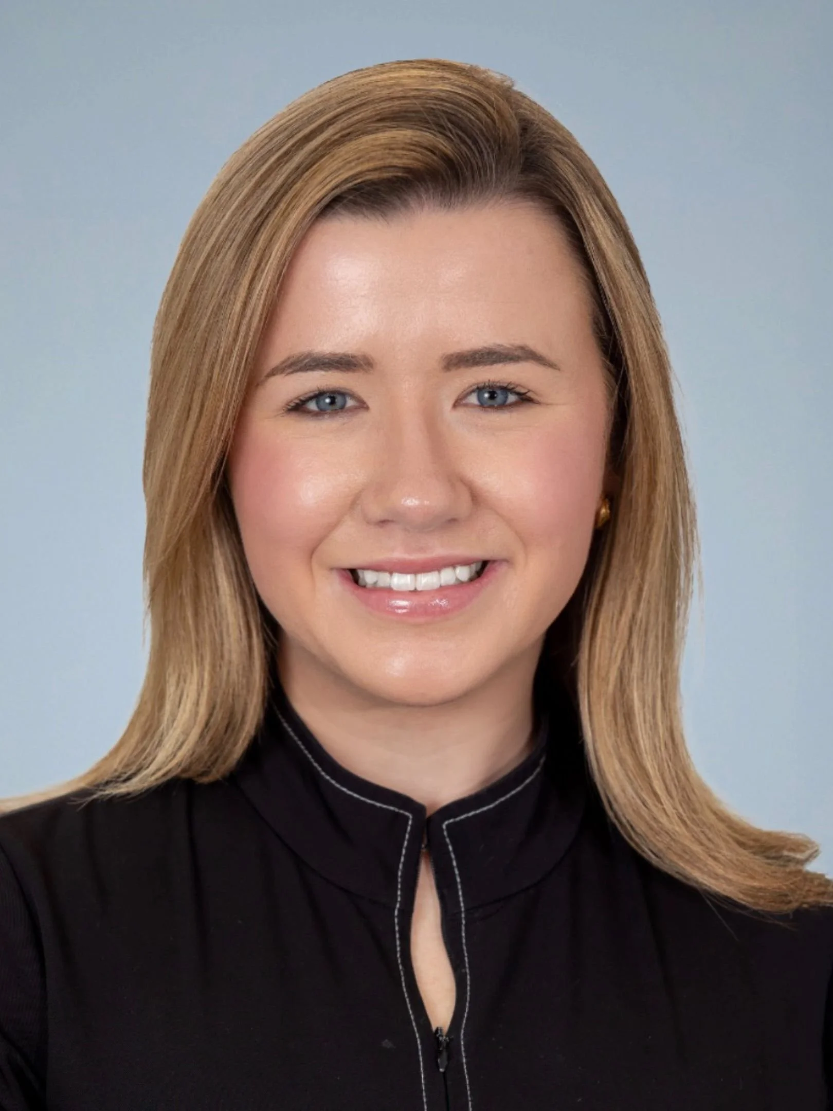 Close-up of a smiling woman with blonde hair, blue eyes, wearing a black top with a zip-up collar, against a light blue background.