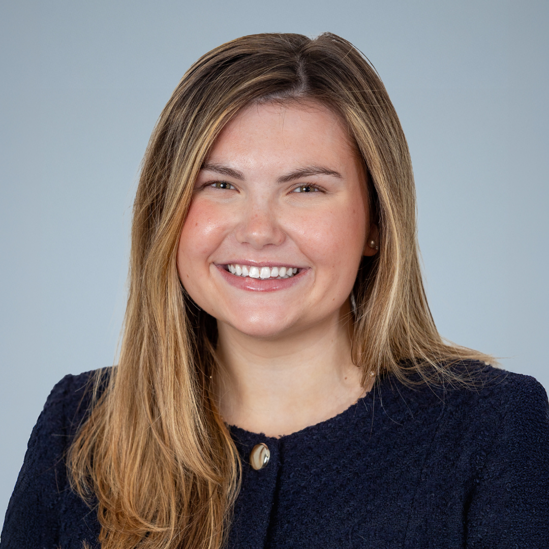 Portrait of a smiling woman with long, light brown hair, wearing a dark blazer with a pearl button, against a neutral background.