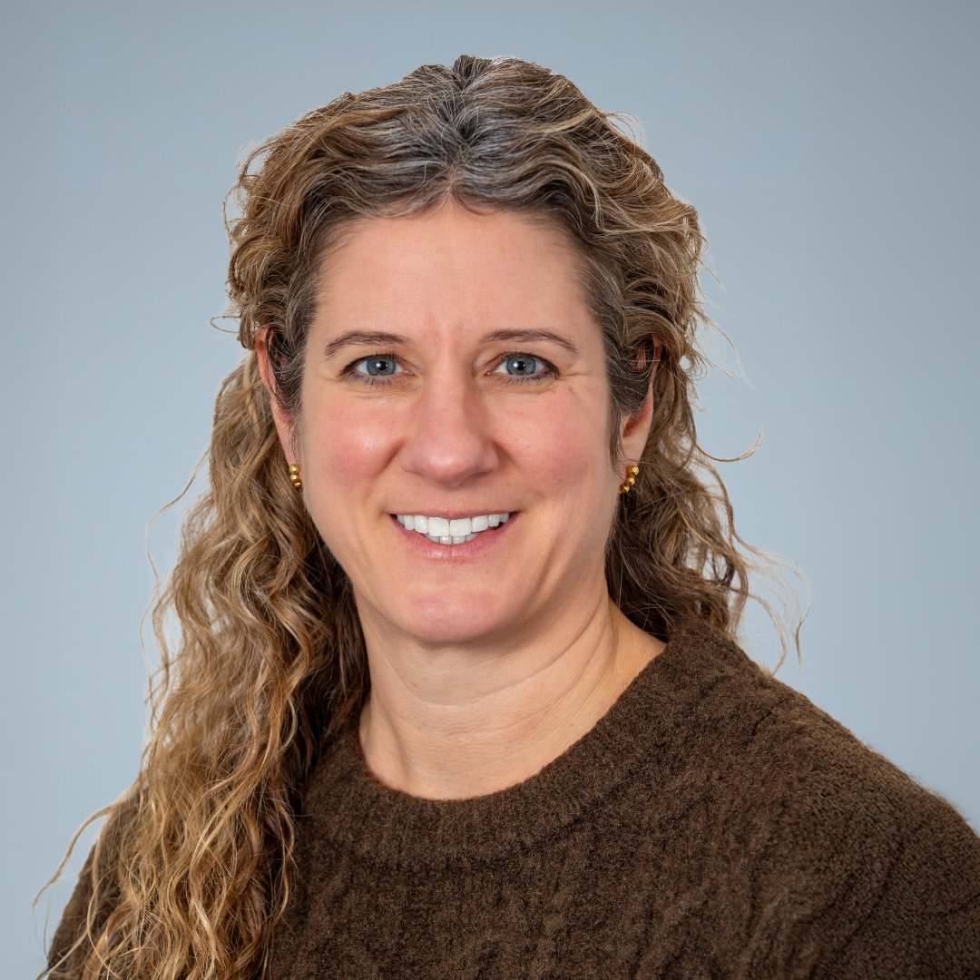 Portrait of a woman with curly, light brown hair and blue eyes, wearing a brown sweater and gold earrings, smiling against a plain gray background.