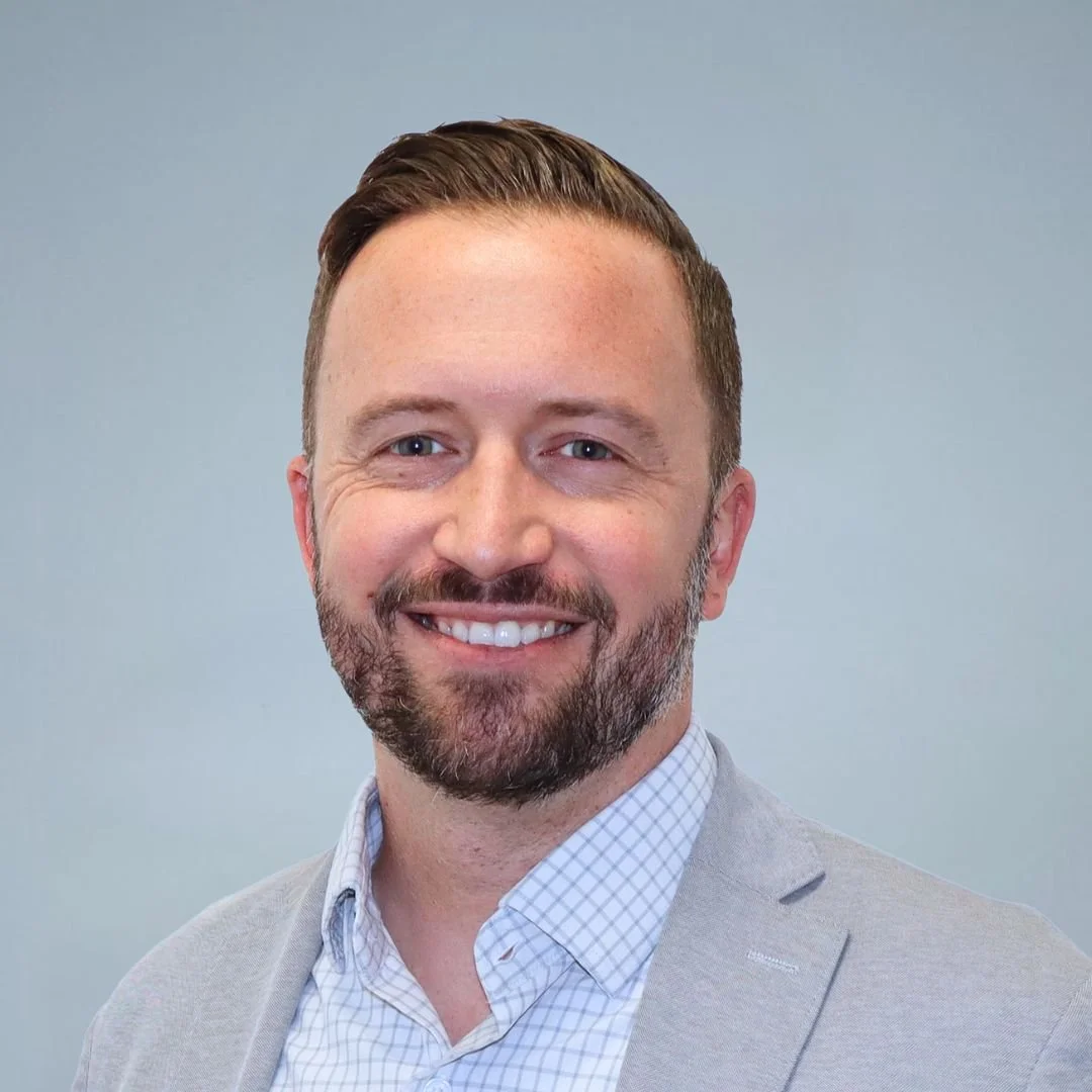 Headshot of a smiling man with short brown hair, beard, wearing a light gray blazer and checked shirt, against a plain gray background.