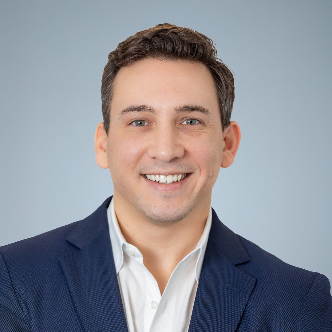 Professional headshot of a man with short dark hair, wearing a navy blazer and white shirt, smiling against a plain light gray background.