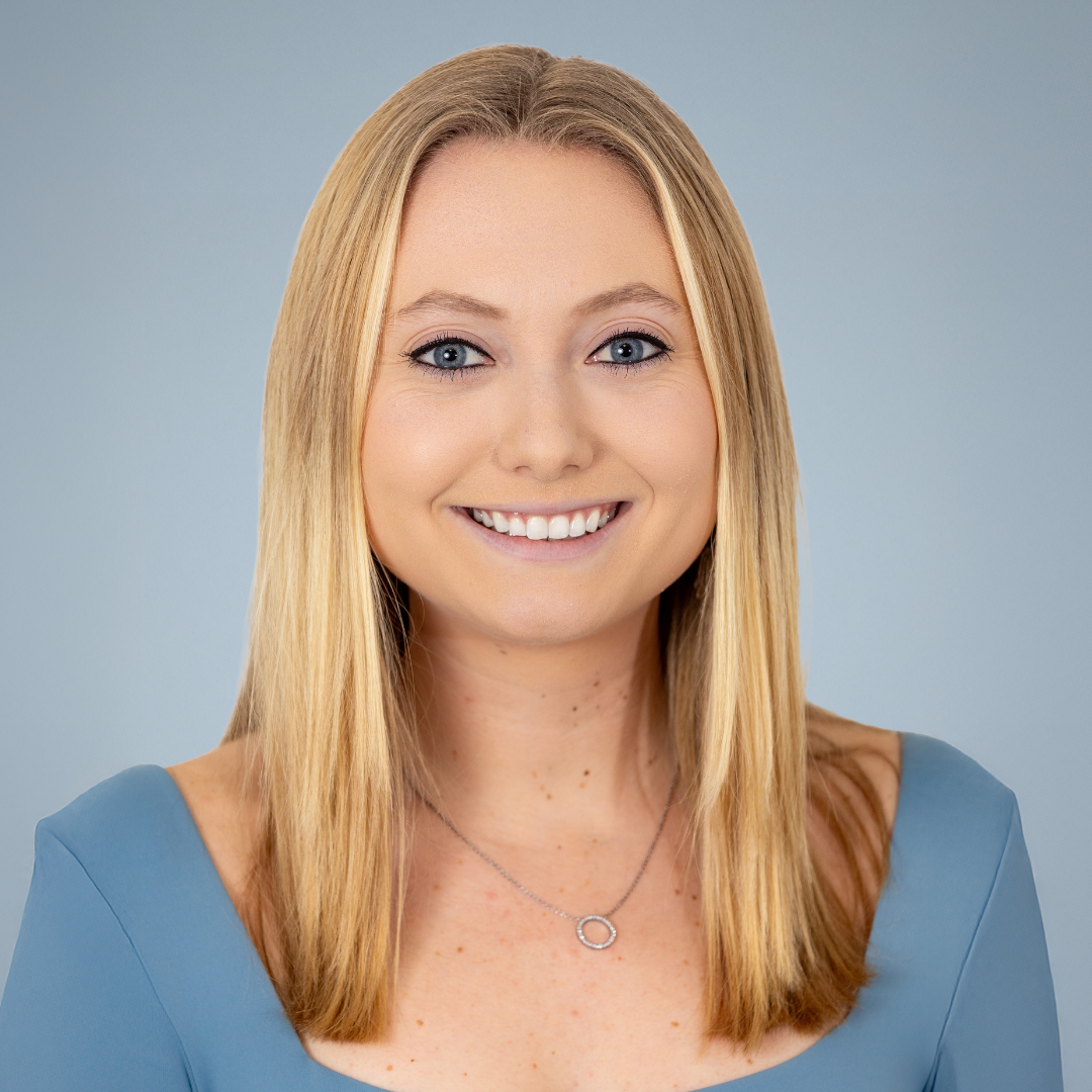 Portrait of a young woman with blonde hair, blue eyes, wearing a light blue top and a silver necklace, smiling against a light grey background.