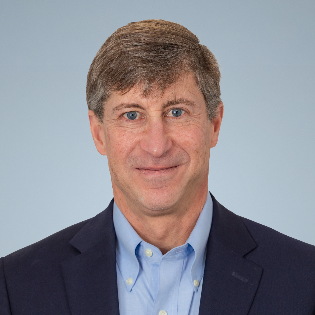 Headshot of a middle-aged man with light brown hair, blue eyes, and a slight smile, wearing a dark suit jacket and light blue collared shirt against a light gray background.