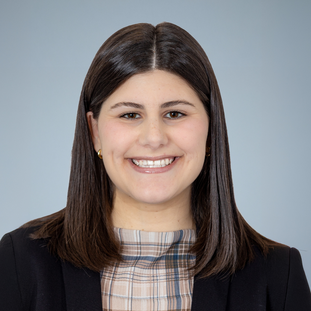 Portrait of a young woman with straight brown hair and brown eyes, smiling, wearing a black blazer and a beige plaid turtleneck.