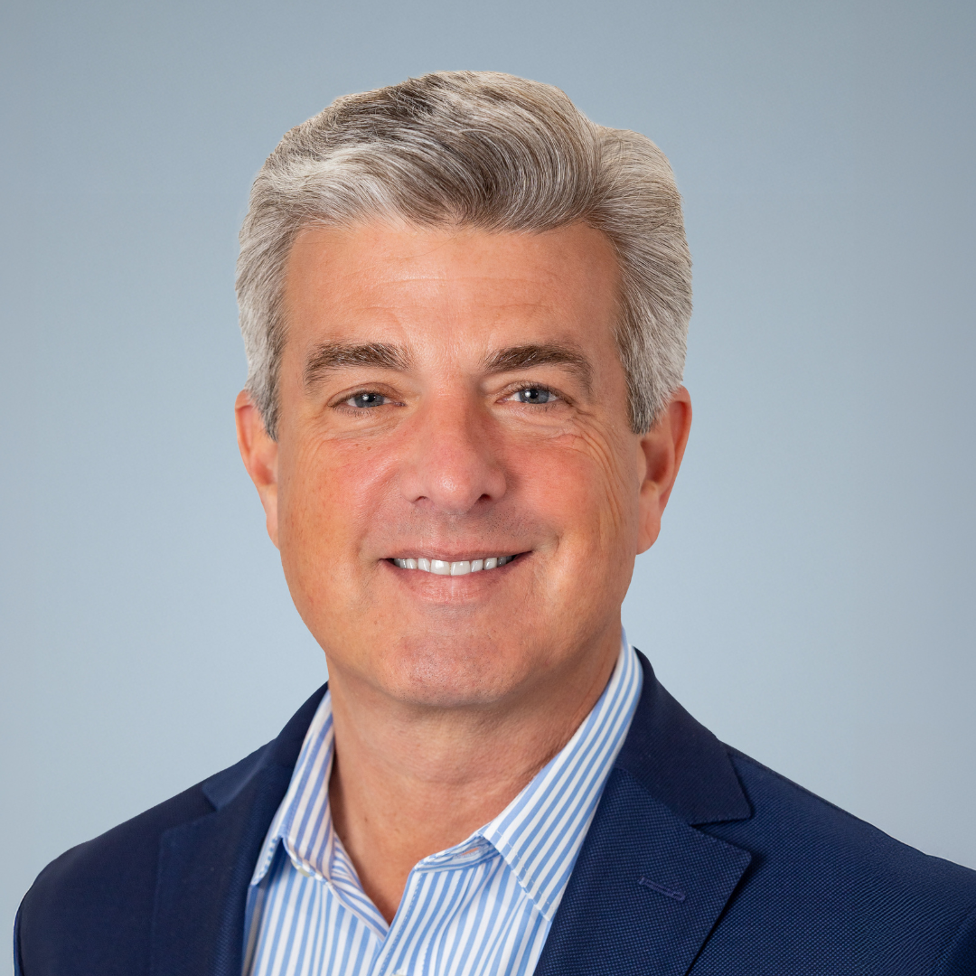 A professional headshot of a smiling middle-aged man with gray hair, wearing a dark blue blazer and a striped dress shirt, against a light blue background.
