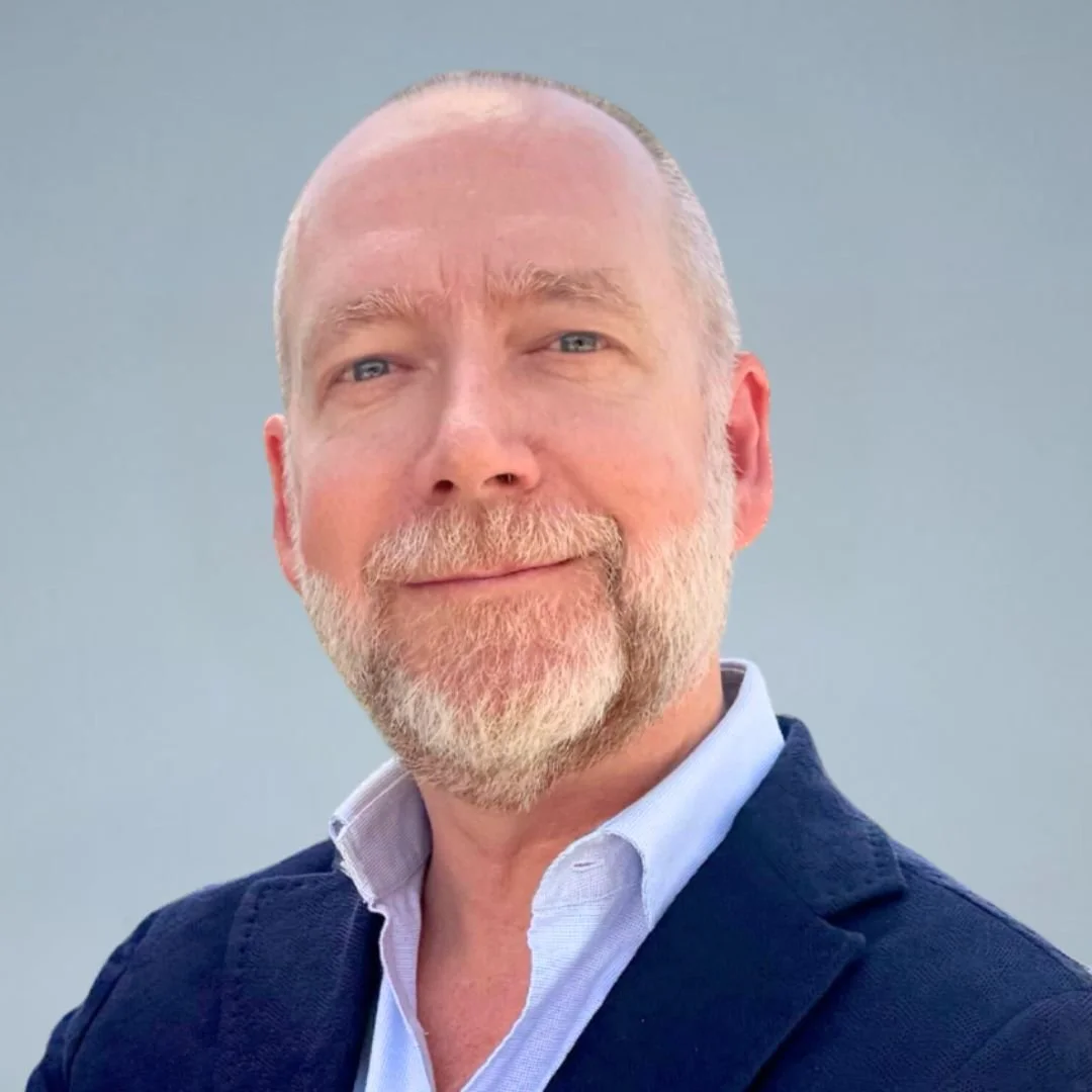 A middle-aged man with a beard and light hair, wearing a navy blazer and white shirt, smiling at the camera against a plain background.
