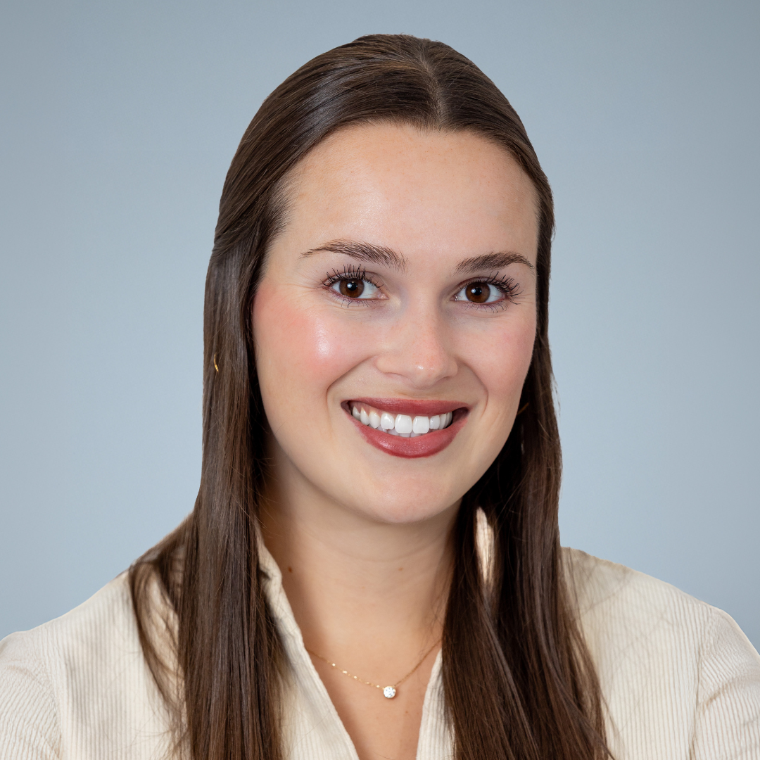 Smiling woman with long brown hair, wearing a cream-colored shirt and a delicate necklace, against a light blue background.