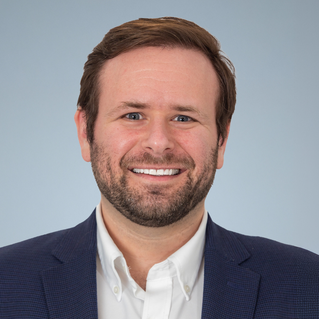 A smiling man with brown hair and a beard, dressed in a white shirt and navy blazer, posing for a professional headshot against a light blue background.