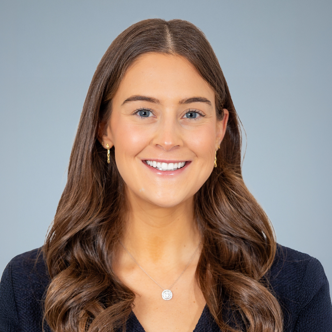 A smiling woman with long brown wavy hair, blue eyes, wearing a black top, small gold earrings, and a silver necklace with a circular pendant, posed against a plain gray background.