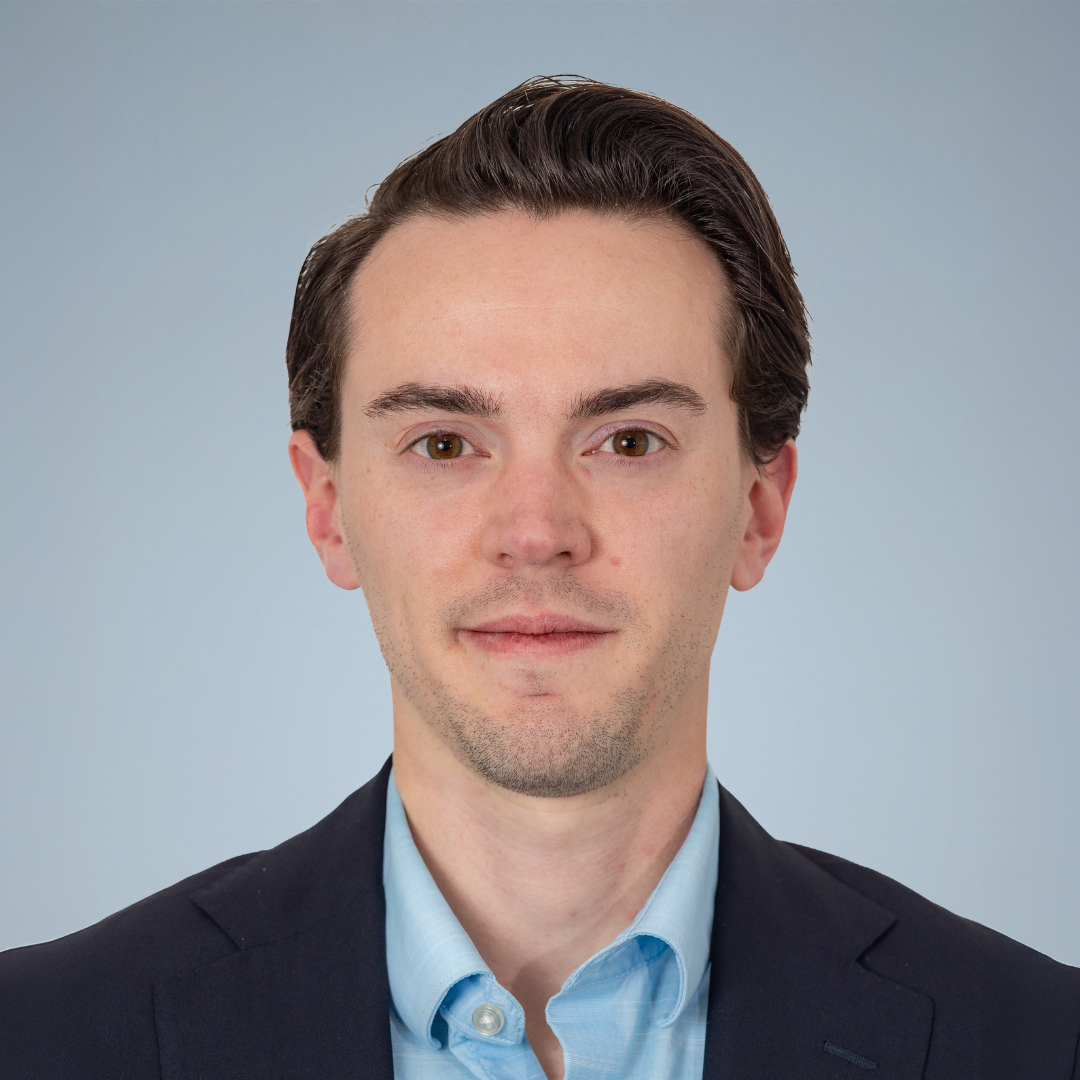 Close-up portrait of a young man with brown hair, wearing a dark suit and light blue shirt, against a plain light background.