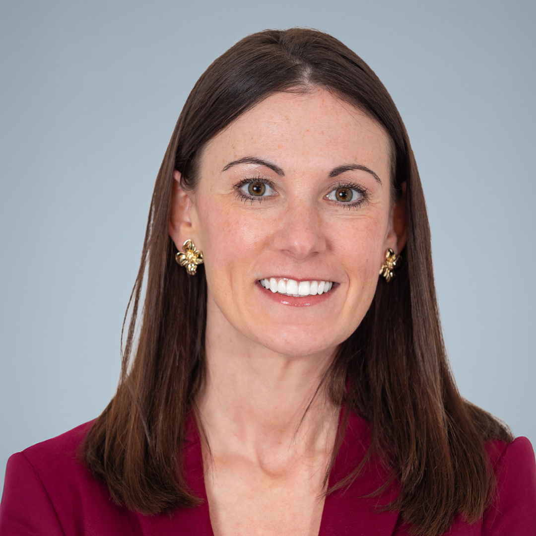 A woman with straight brown hair, wearing gold earrings and a burgundy blazer, smiling against a light gray background.
