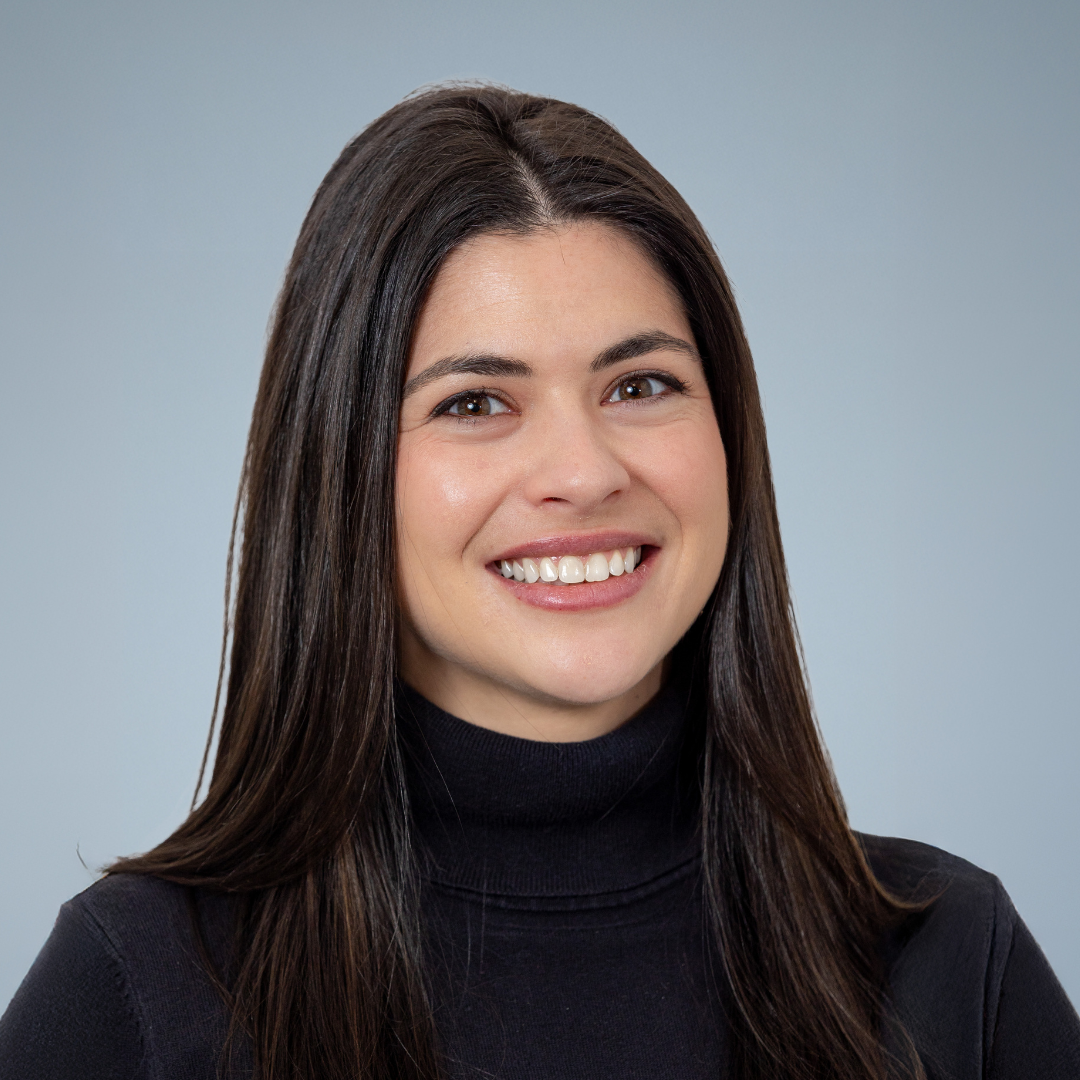 A woman with straight dark hair smiling, wearing a black turtleneck, against a plain grey background.