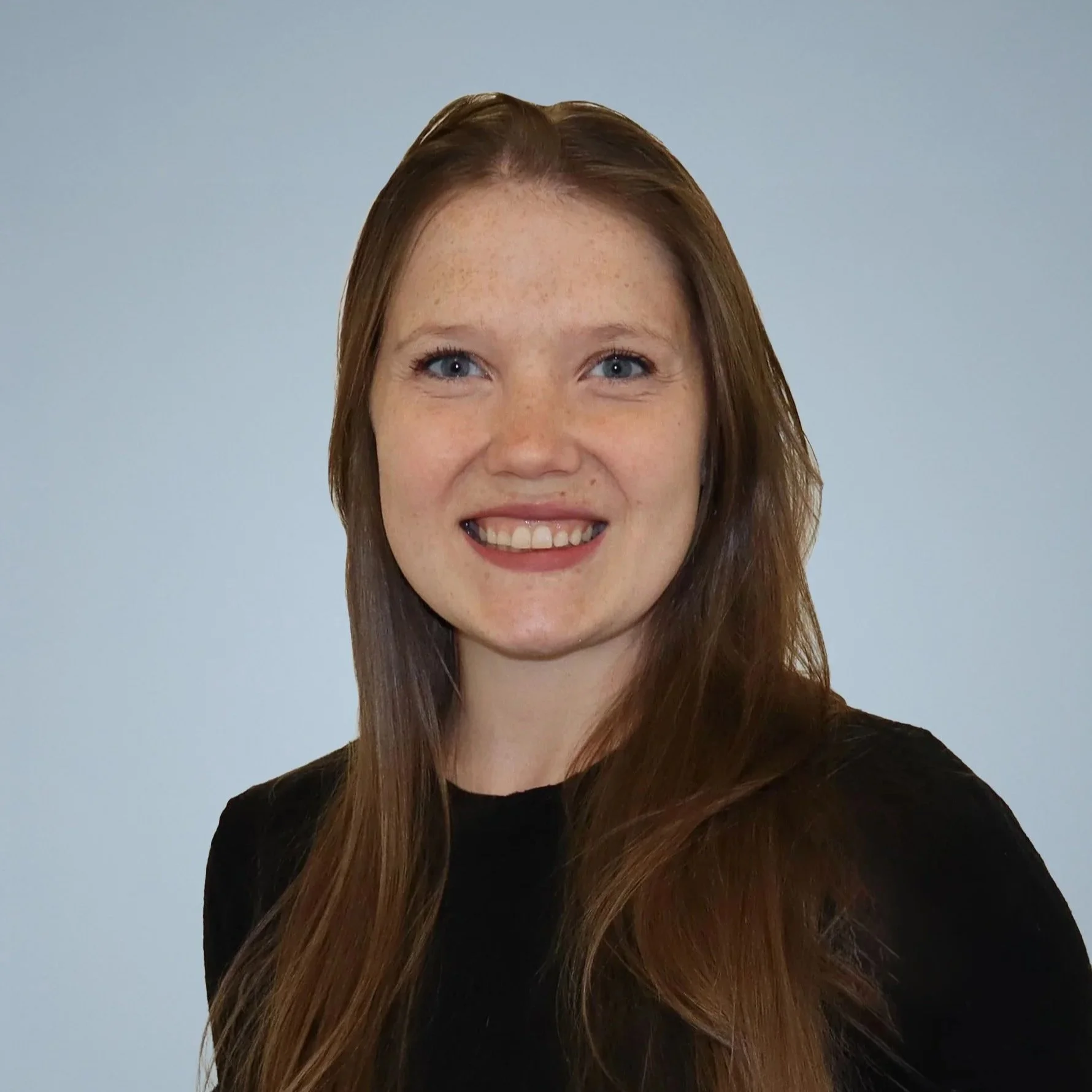 A young woman with long brown hair, blue eyes, freckles, and a wide smile wearing a black shirt, posing against a plain light blue background.